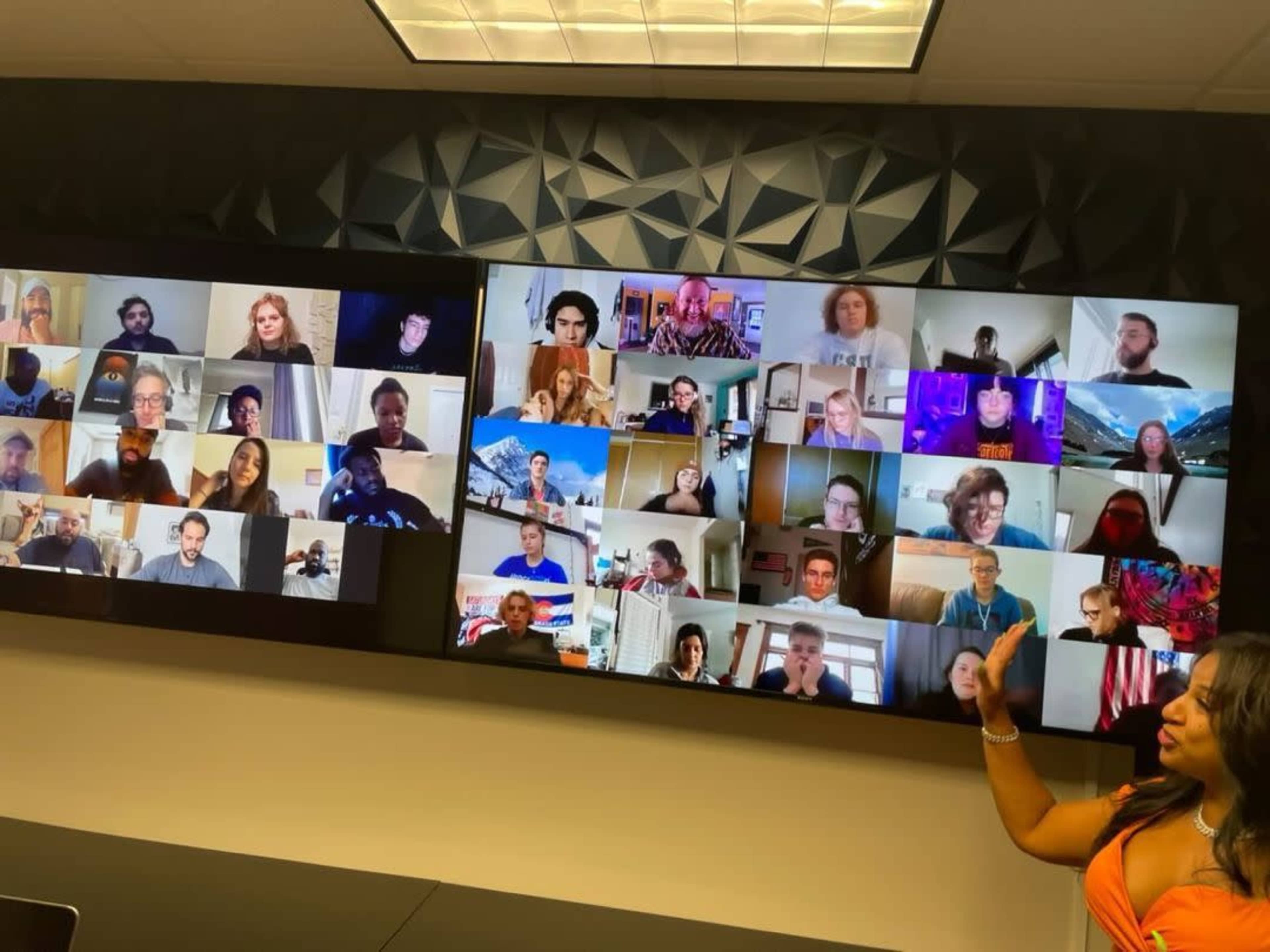 A person gestures towards multiple screens displaying a large group of people participating in a virtual meeting.