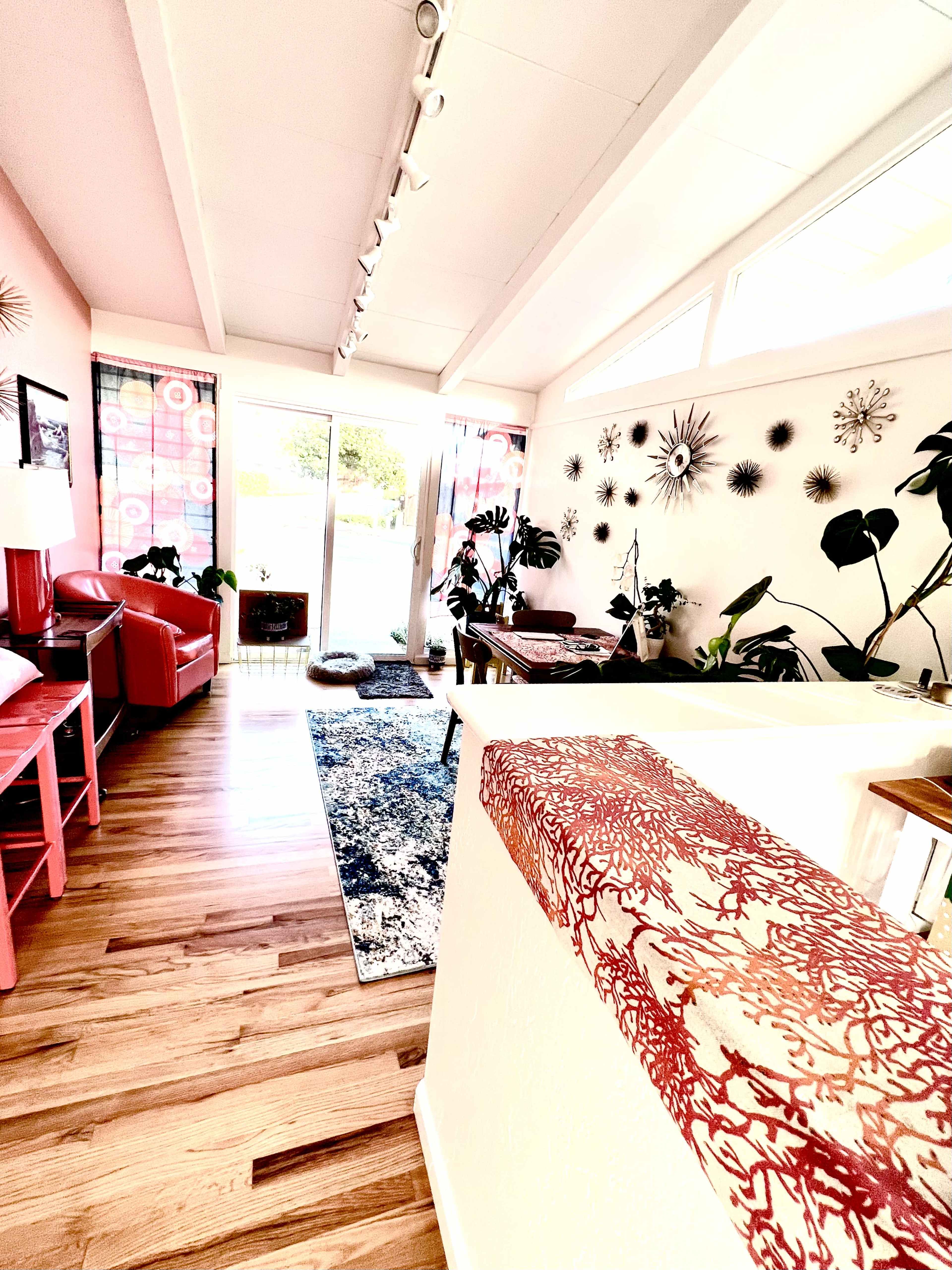 A bright living space featuring hardwood floors, a red lounge chair, and large windows with plants and colorful decorations along the walls.