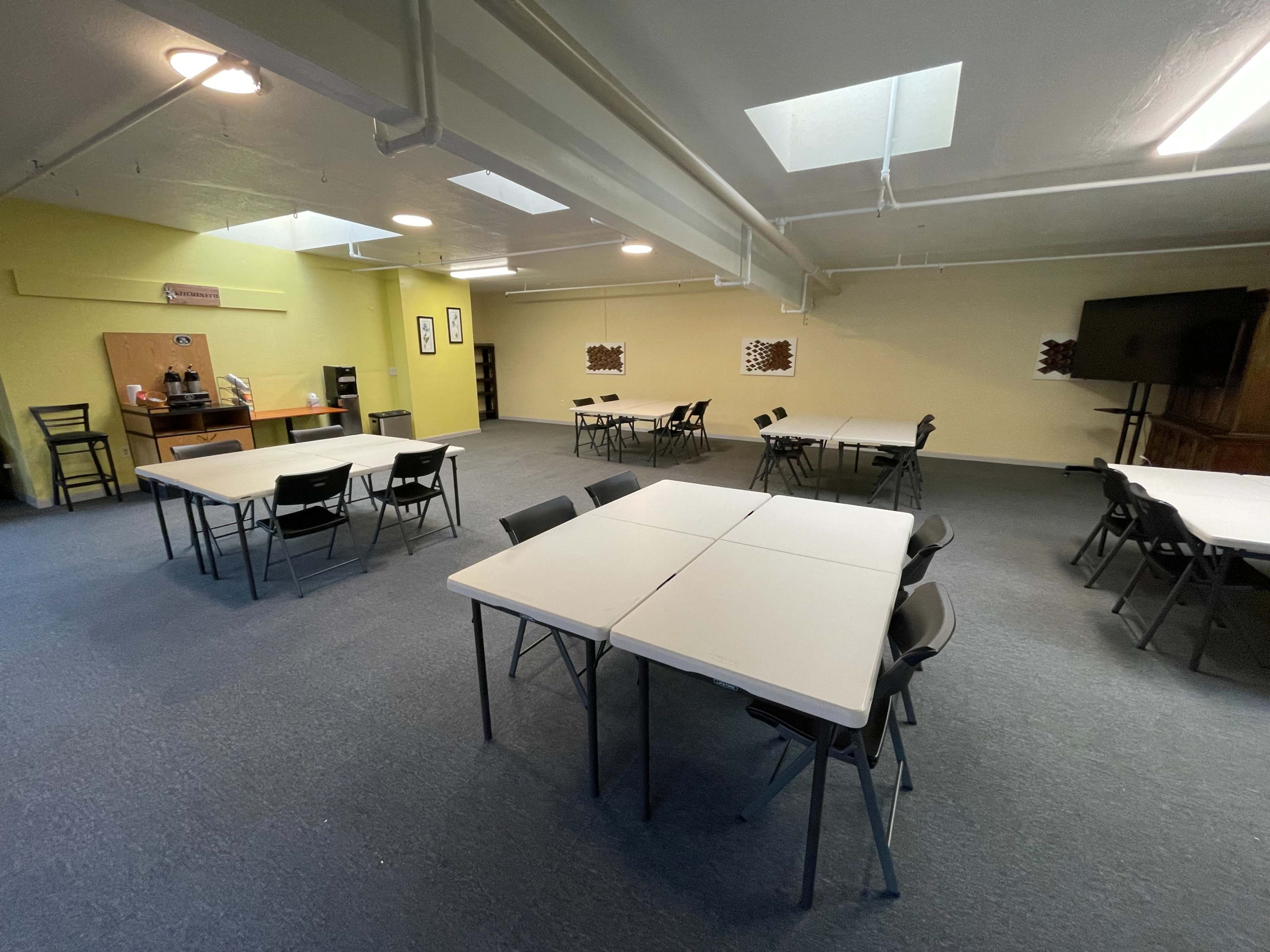A spacious room with several tables and chairs arranged for meetings or events, featuring a coffee station in one corner.