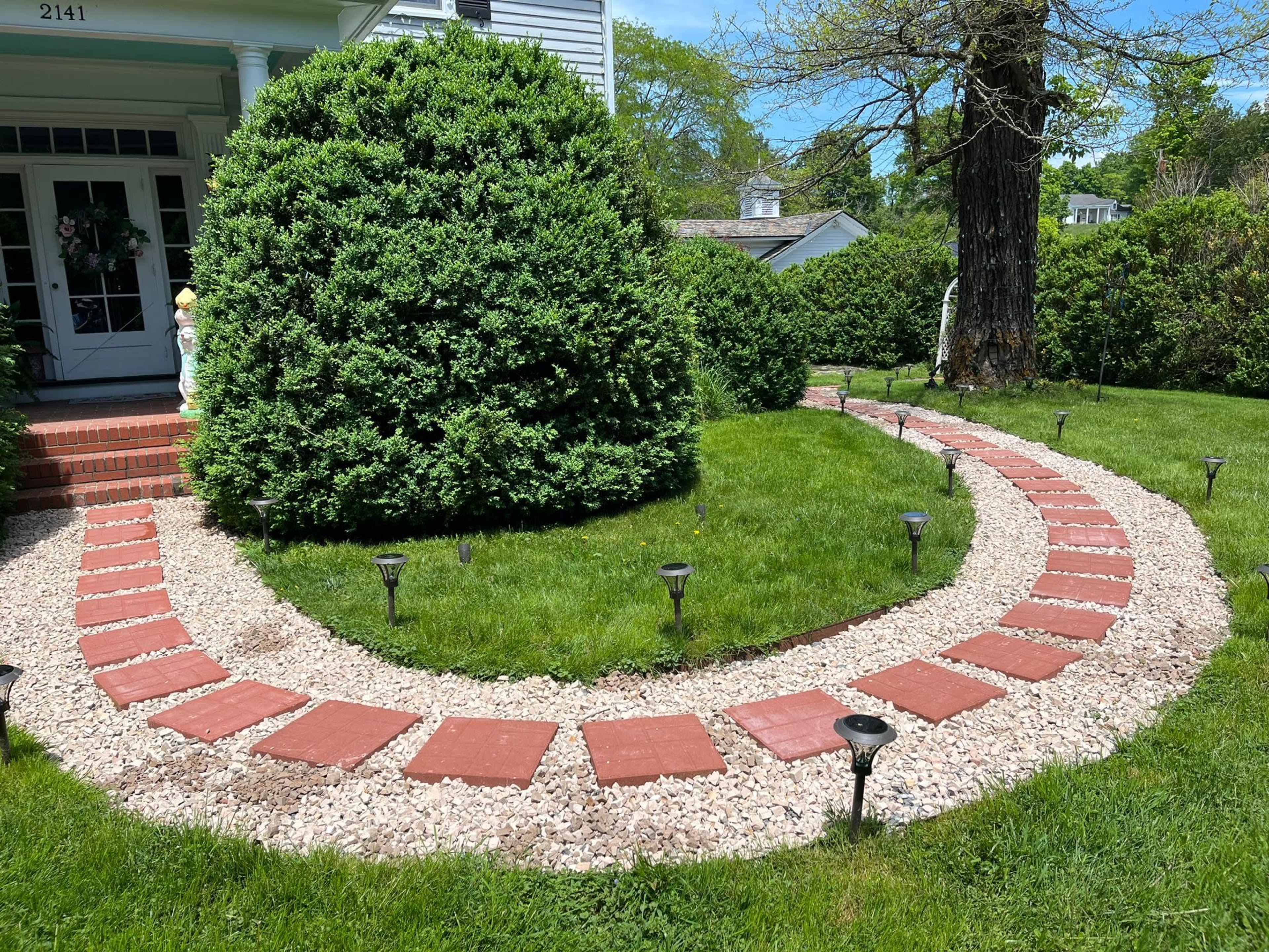 A landscaped garden pathway made of red bricks and gravel, winding through a grassy area with bushes and garden lights.