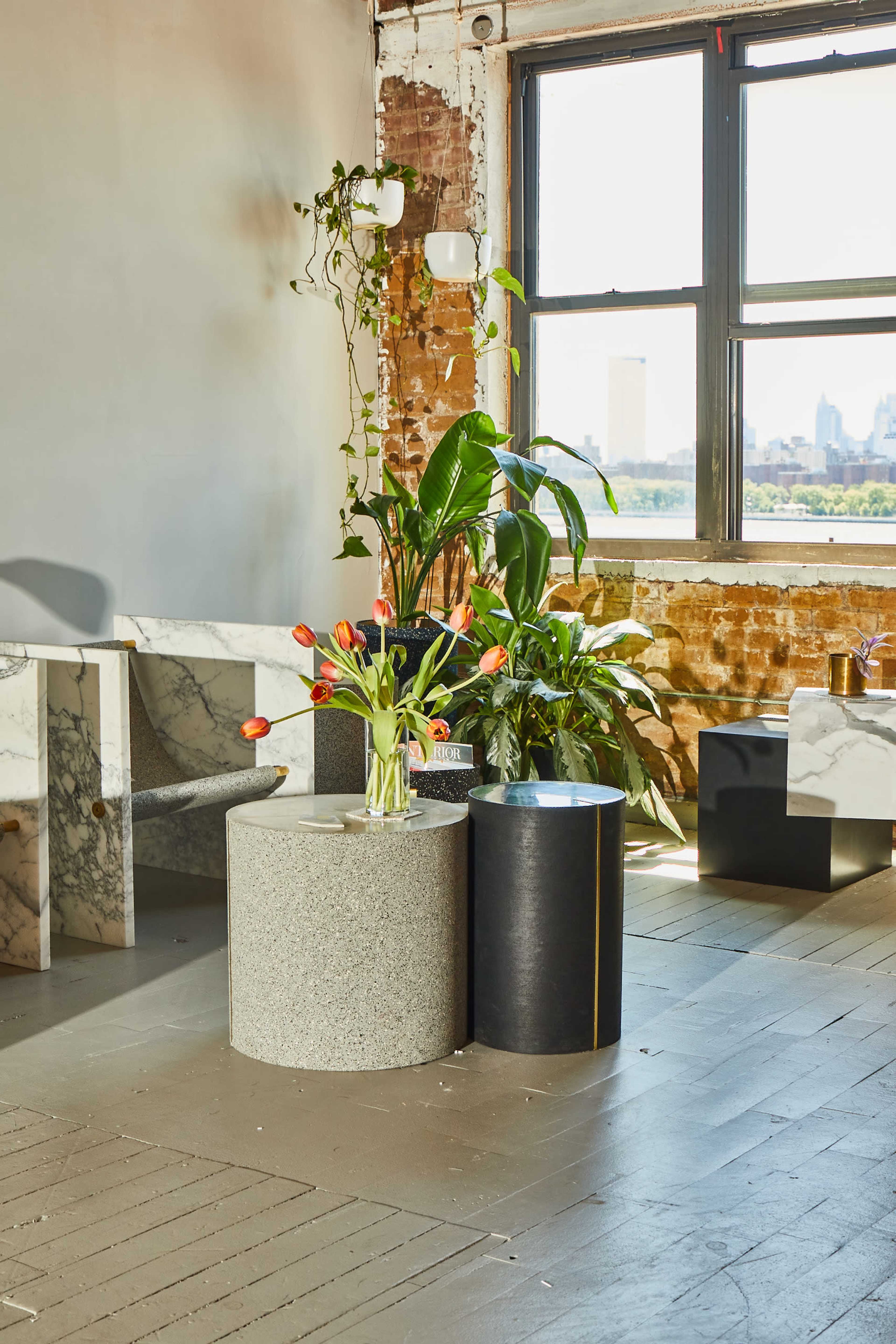 A bright interior with a large window, featuring various plants and sleek, cylindrical tables made of stone, set against a backdrop of exposed brick walls.