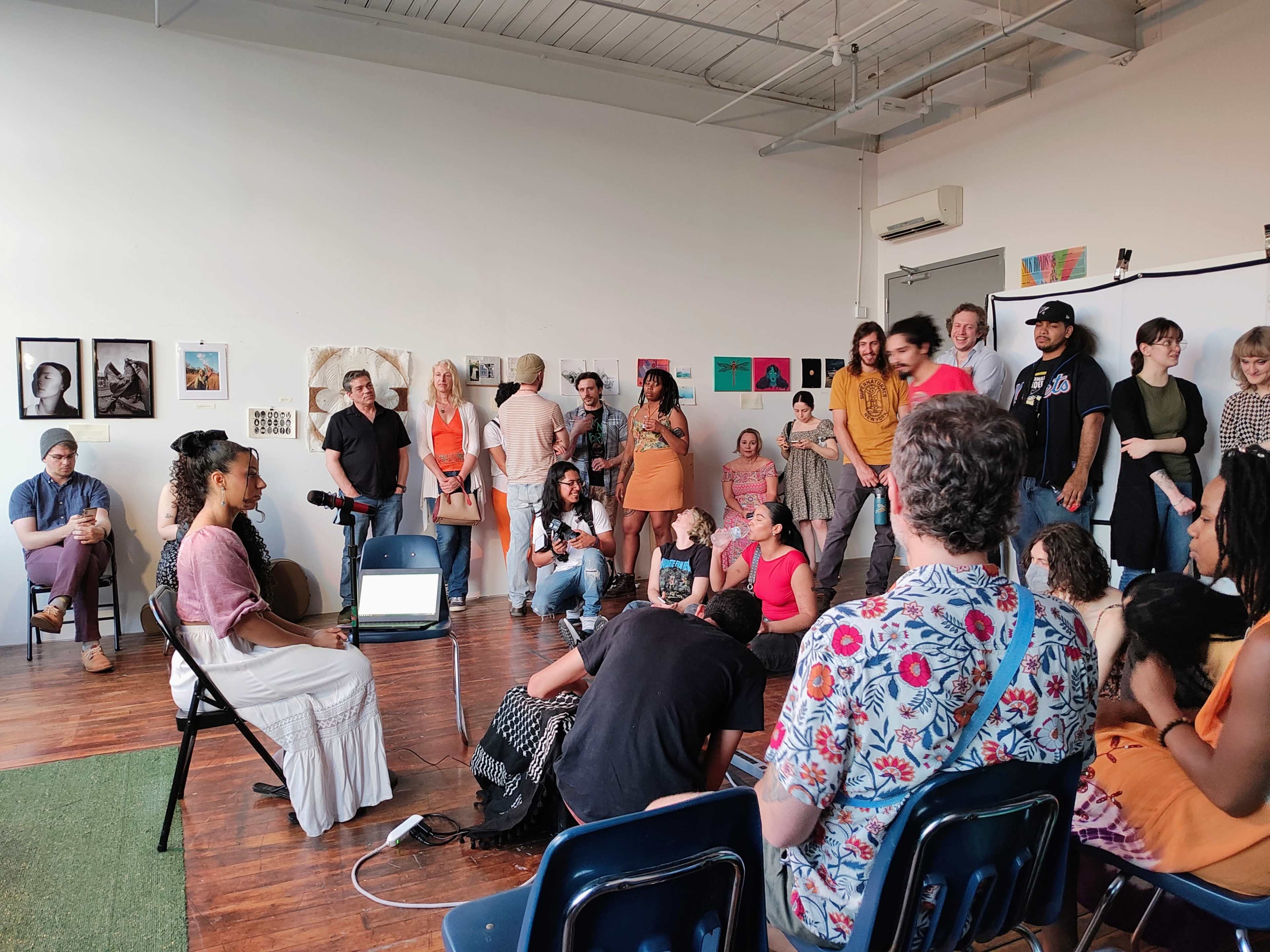 A diverse group of people gathers in a bright room for an event, with some seated and others standing near a speaker in front of a projection screen.