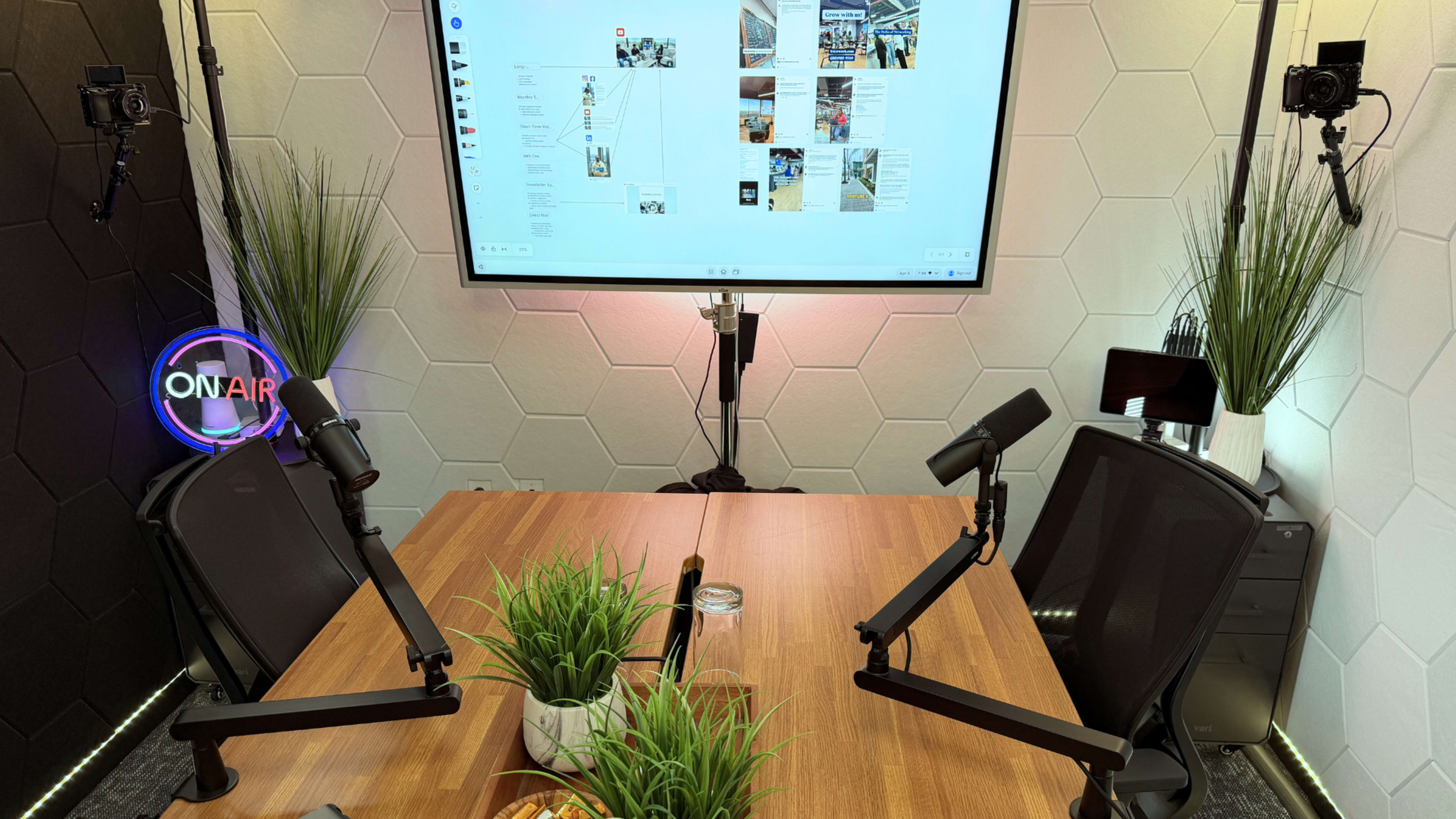 The image shows a modern recording studio setup with a wooden table, two black chairs, a large screen displaying a digital interface, and plants near the table.
