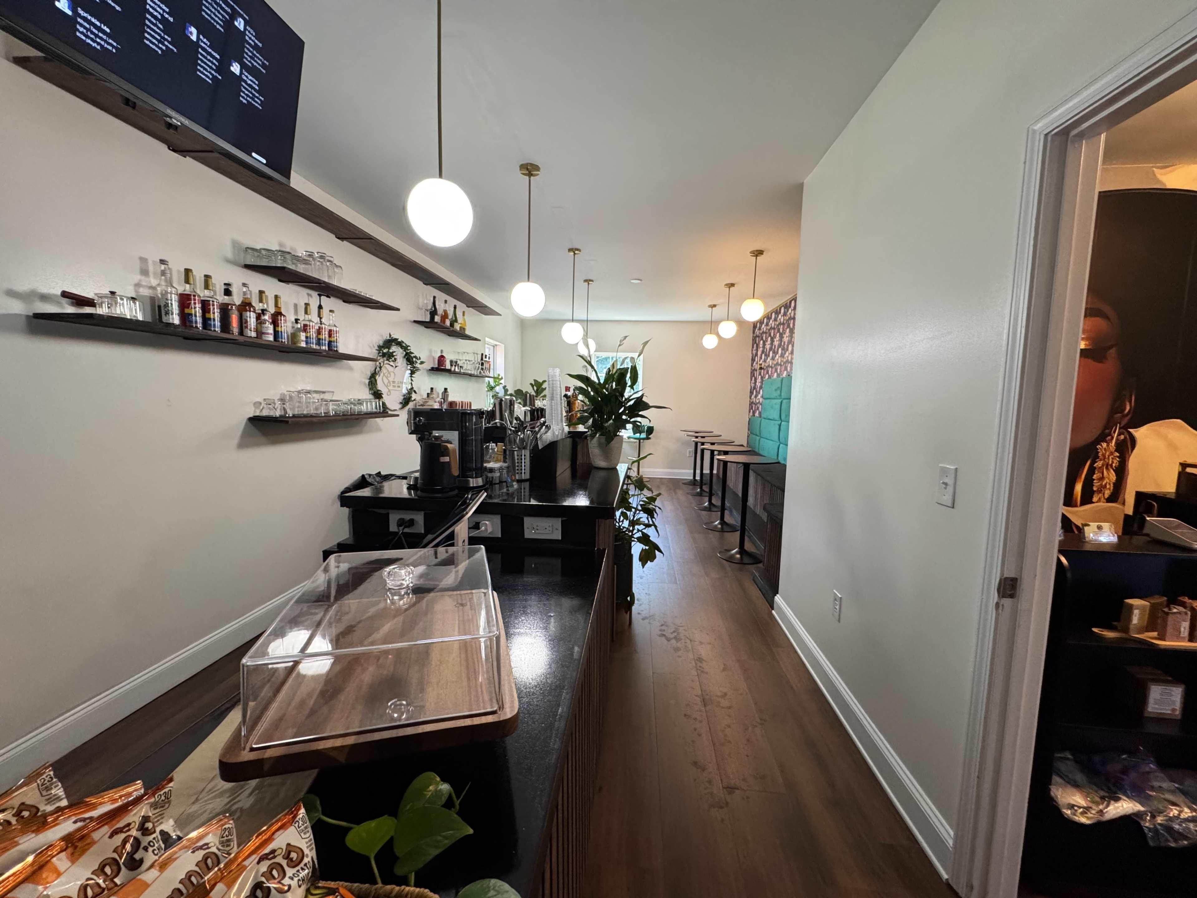 The image depicts a modern coffee shop interior with a long bar area, shelving displaying various beverage options, and seating along the wall.