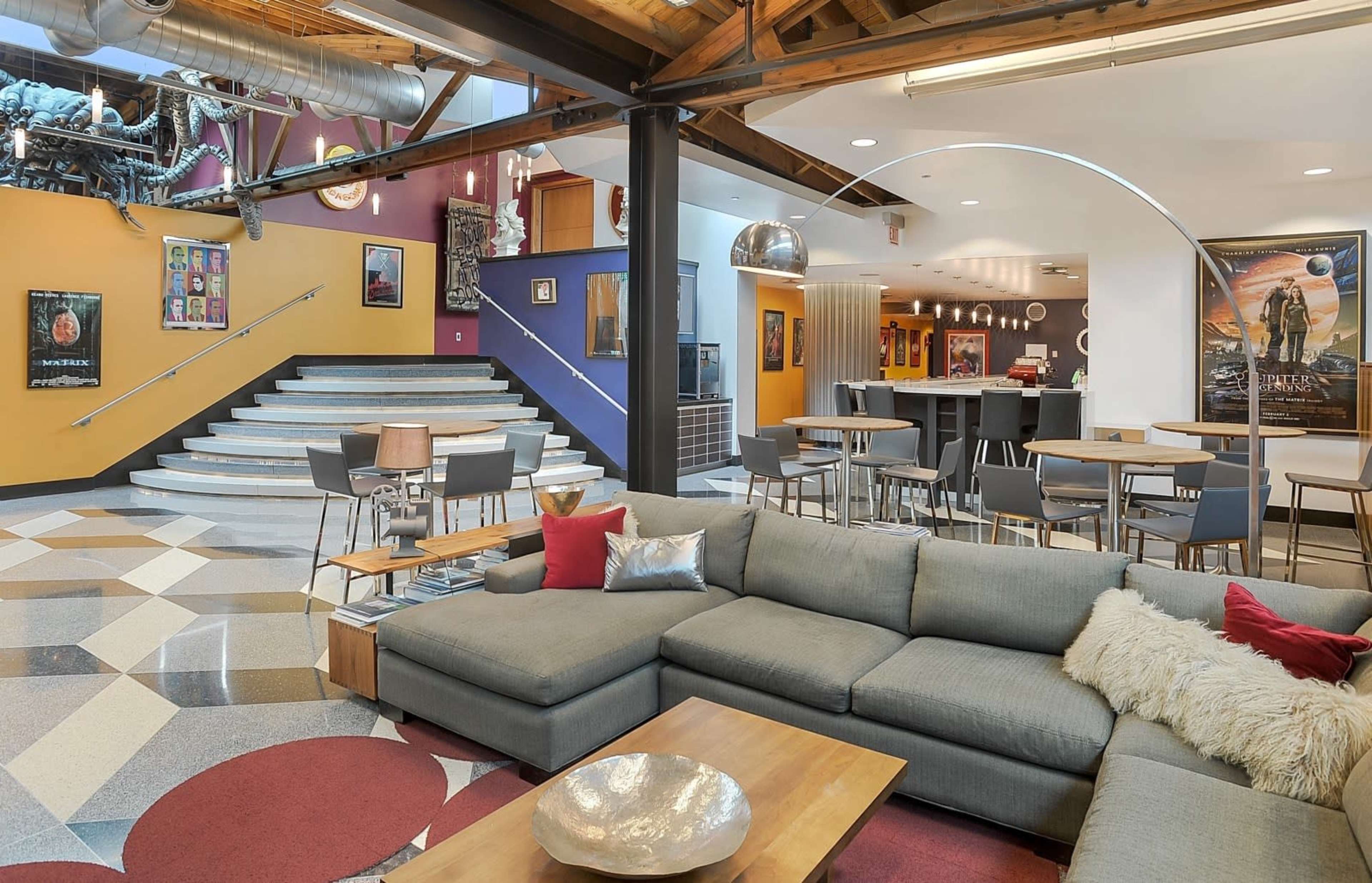 A modern open-concept living space with a large gray sectional sofa, a coffee table, and a staircase leading to an upper level, surrounded by colorful walls and various artworks.