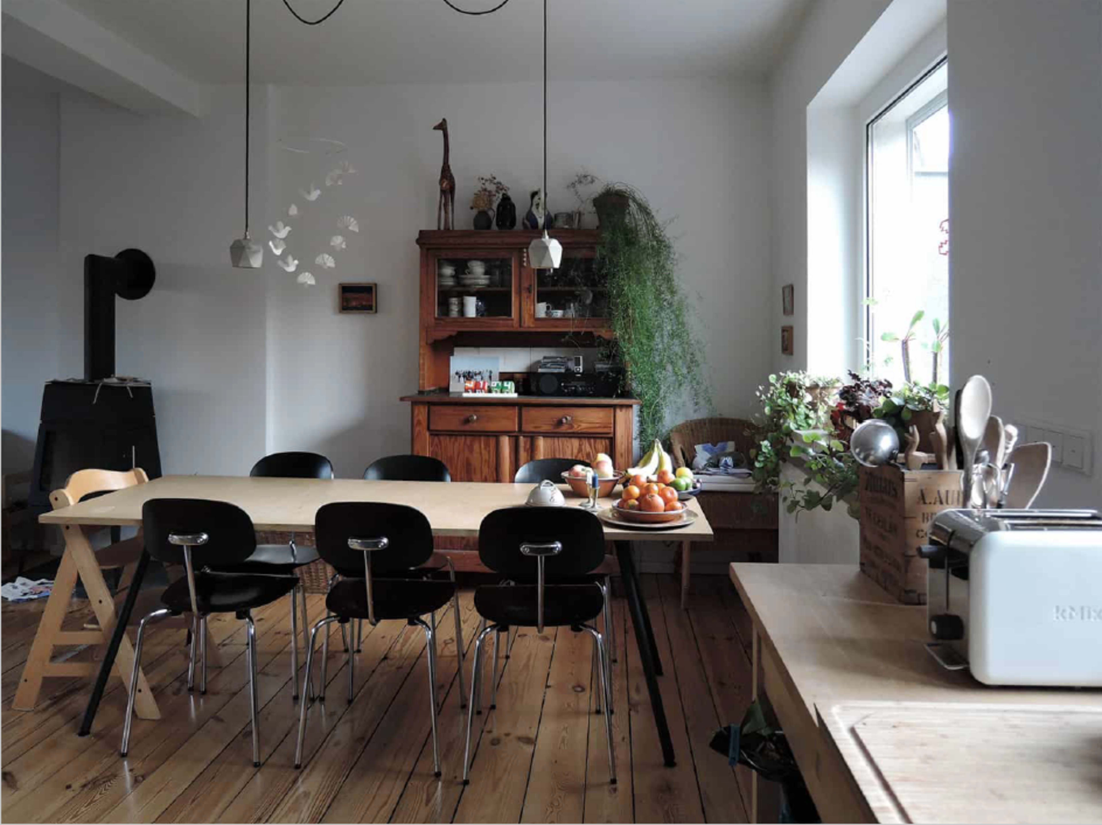 A modern kitchen with a wooden dining table, six black chairs, and a sideboard filled with kitchenware, surrounded by plants and natural light.