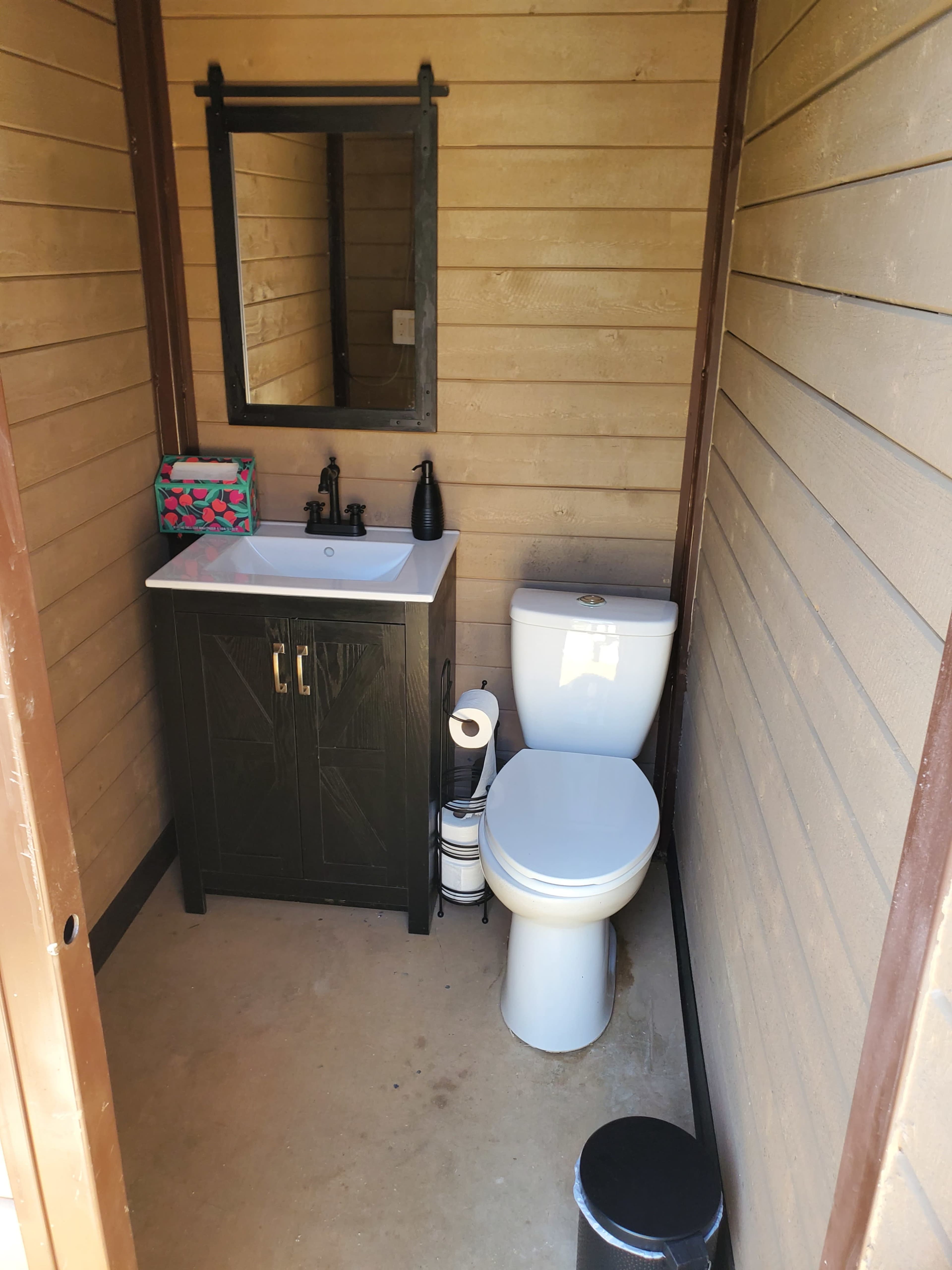 The image shows a small bathroom with a sink, mirror, toilet, and a waste bin, all enclosed in wood-paneled walls.
