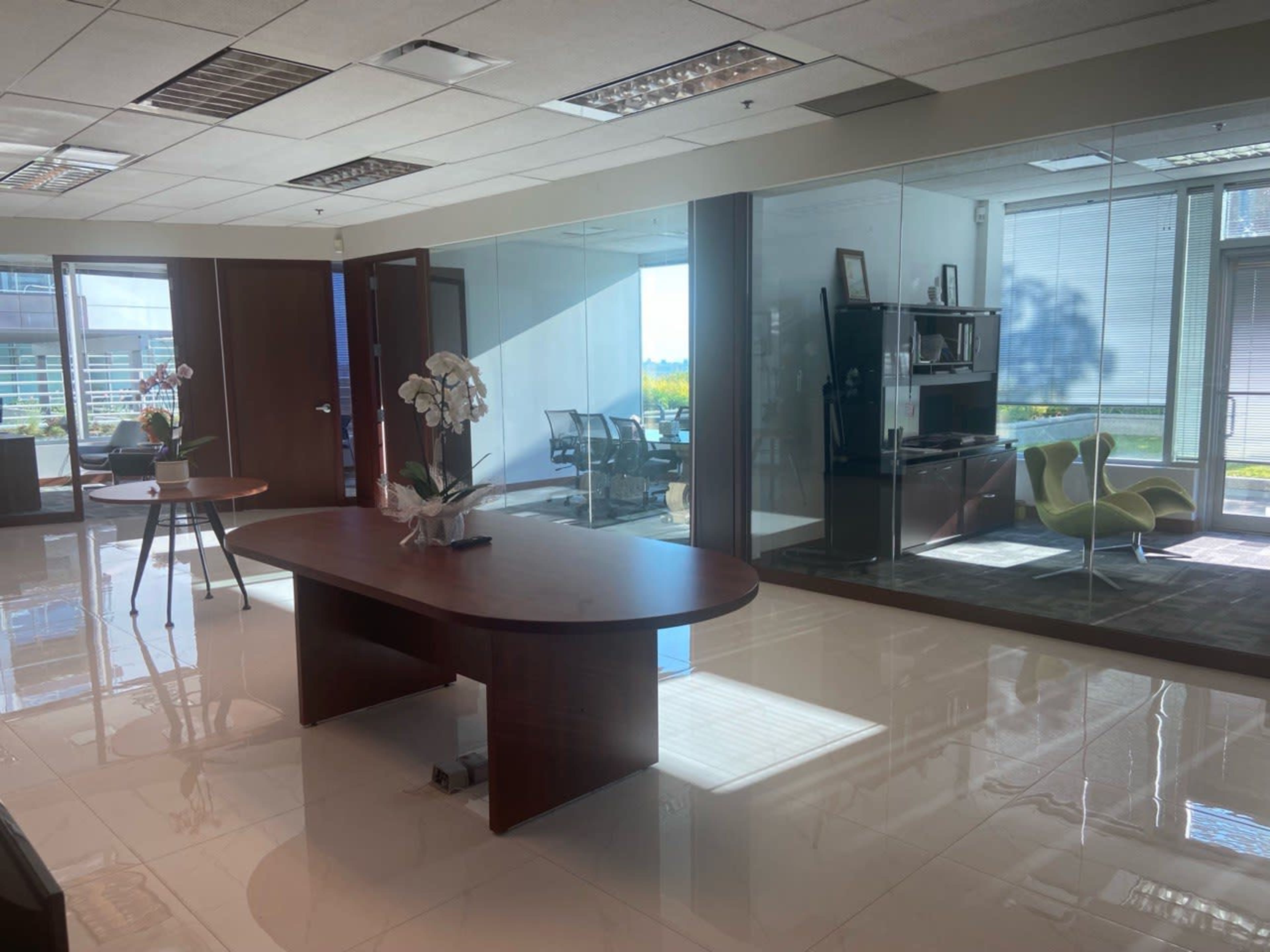 An office space featuring a central wooden table, glass walls, and modern furniture, with natural light streaming in through large windows.
