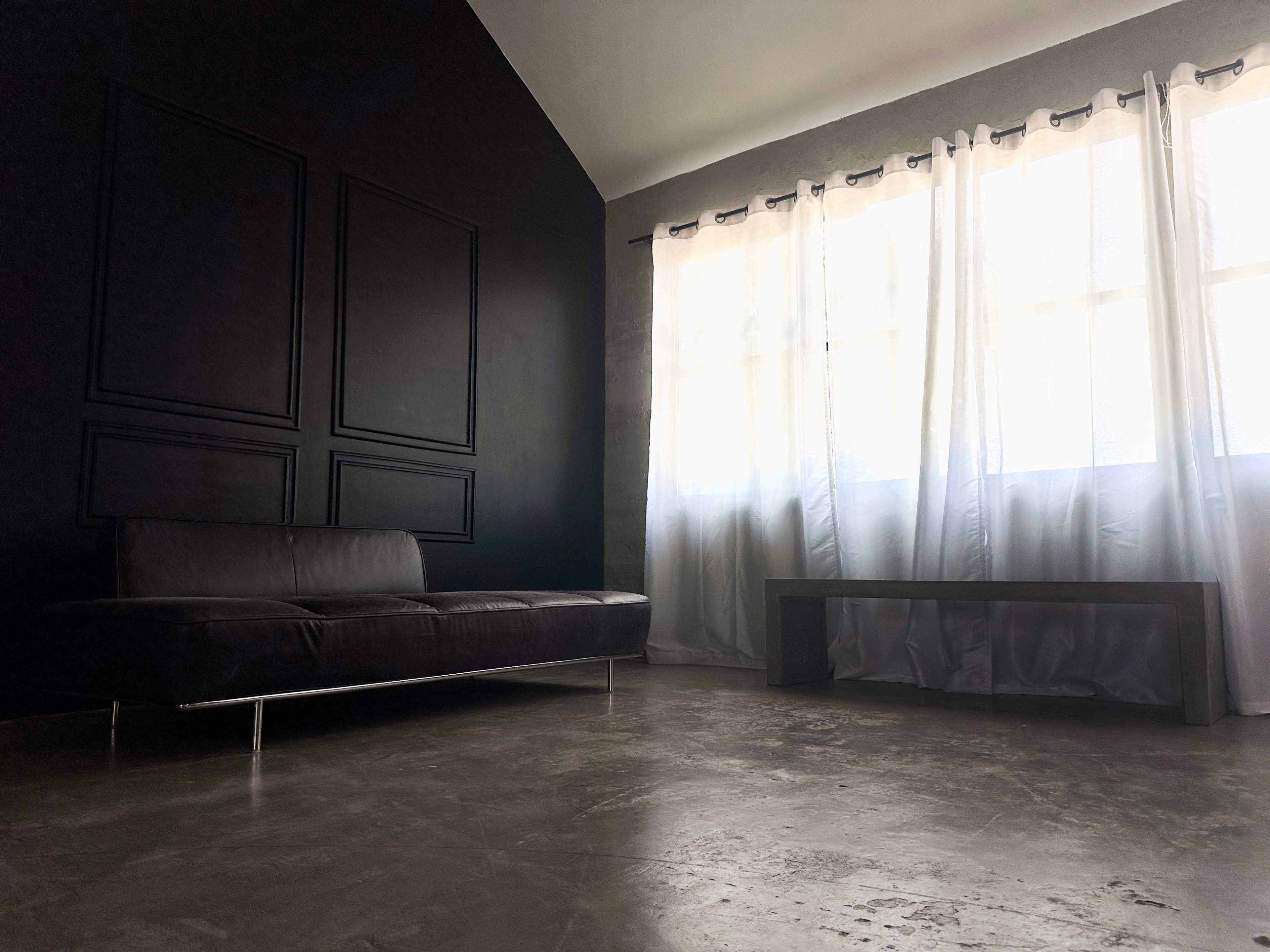 A minimalist living space featuring a black wall, a dark sofa, and large white curtains covering a window.