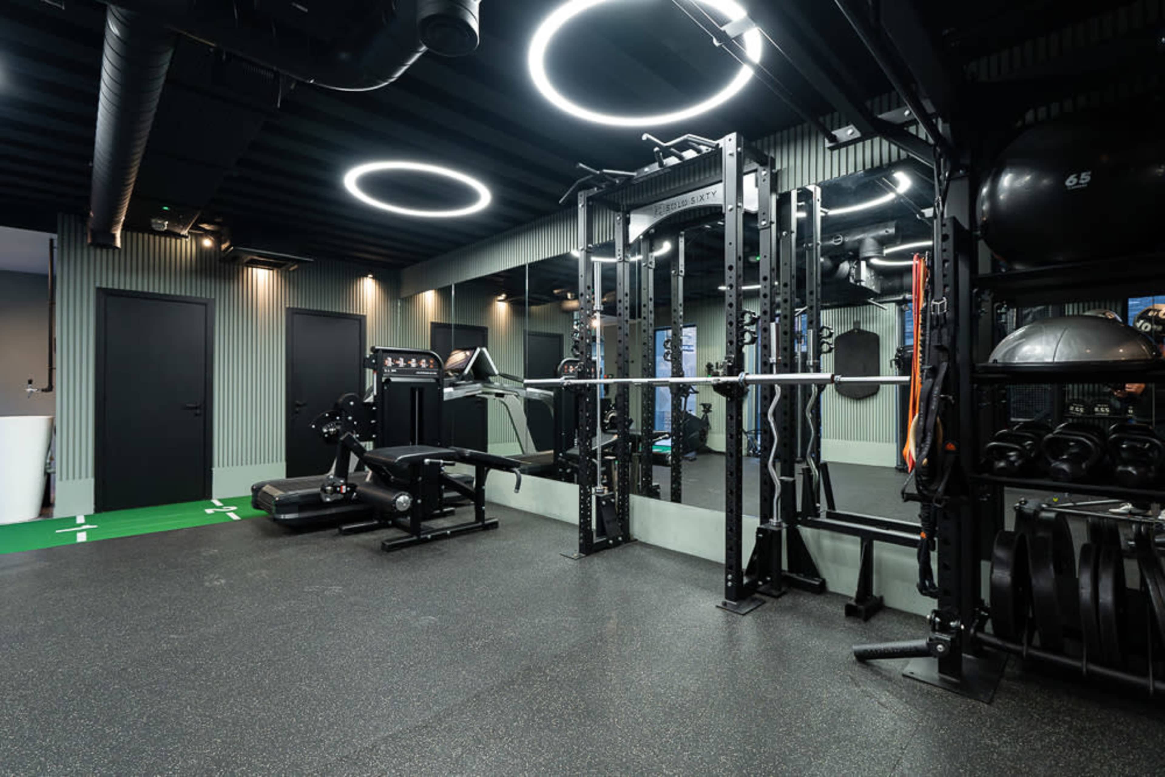The image shows a modern gym interior featuring exercise equipment, including a weightlifting station, cardio machines, and various fitness accessories.