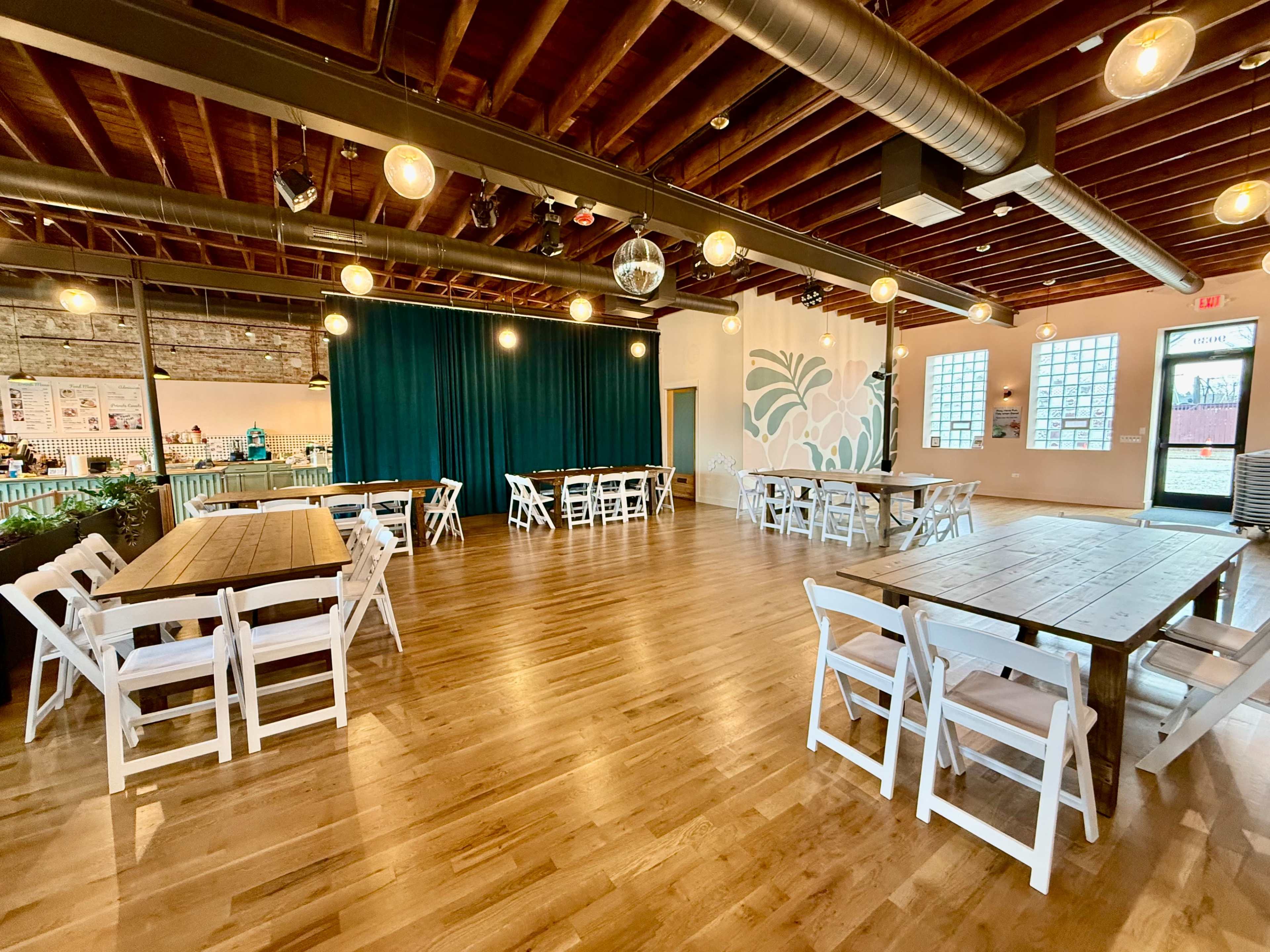 The image shows a spacious dining area with wooden floors, several white folding tables, and green curtains in the background.