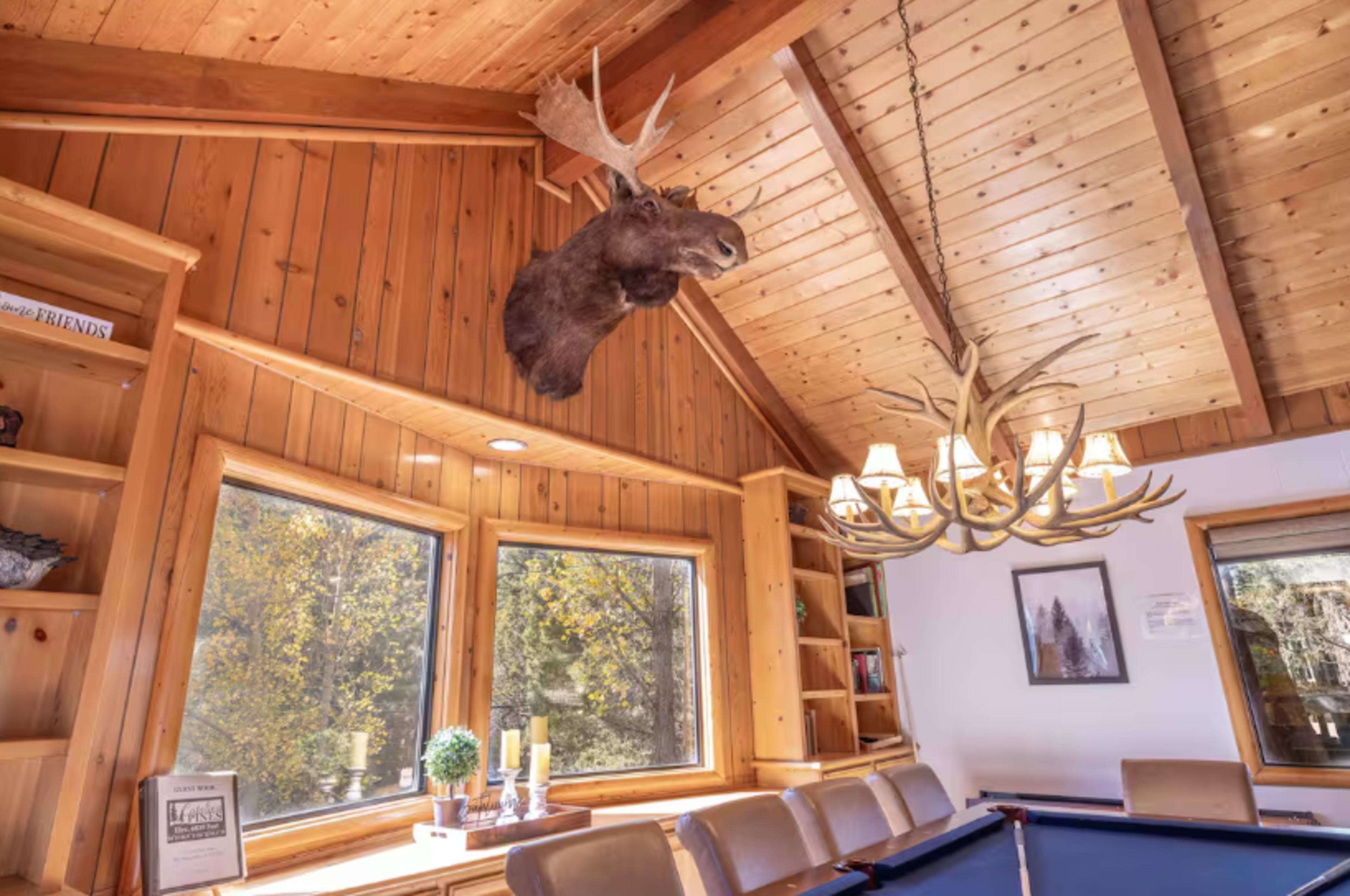 A mounted moose head hangs above a seating area with large windows, and a chandelier made of antlers complements the wooden interior.