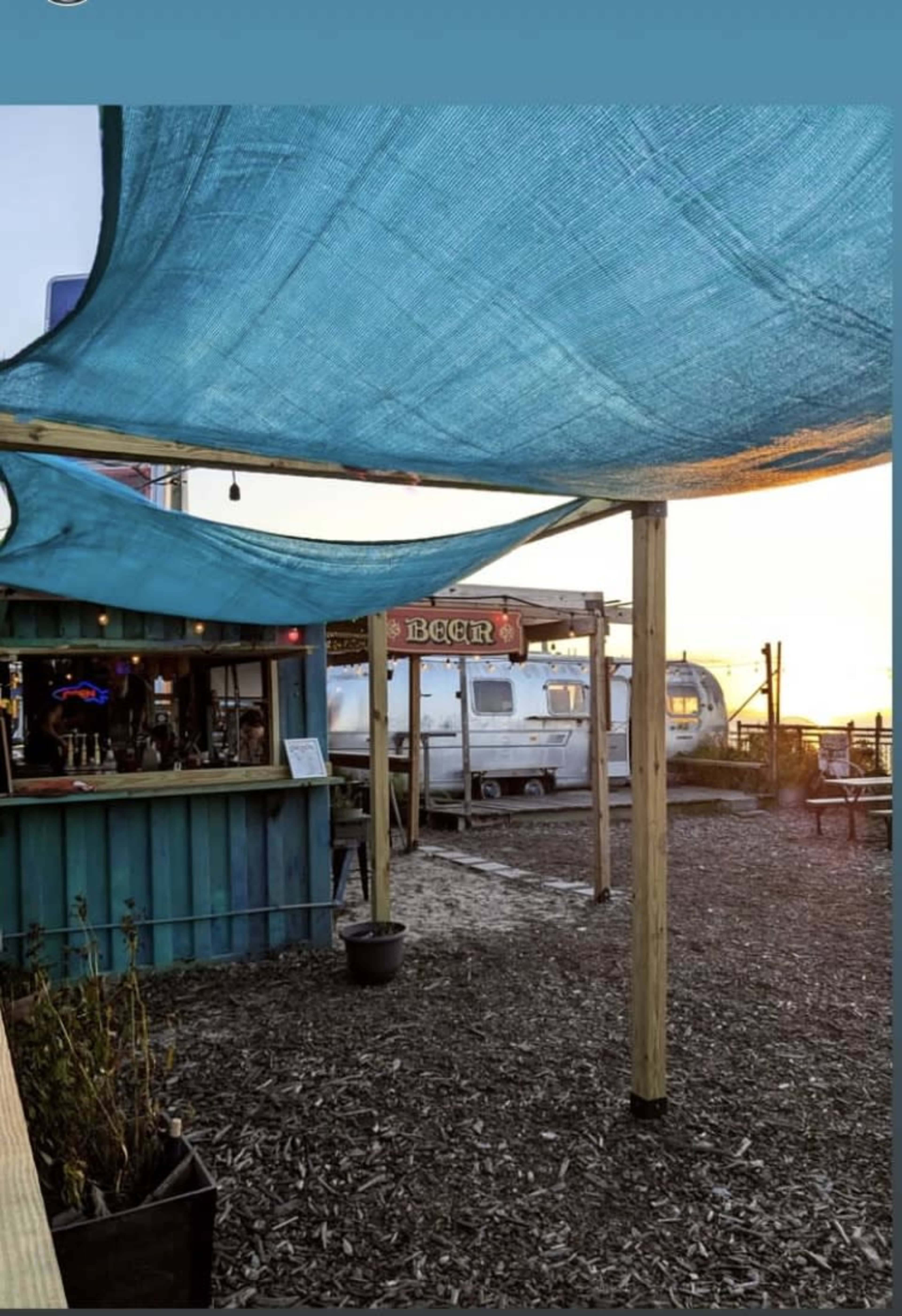 The scene shows a bar with a blue canopy and vintage trailers in the background, set along a waterfront area during sunset.