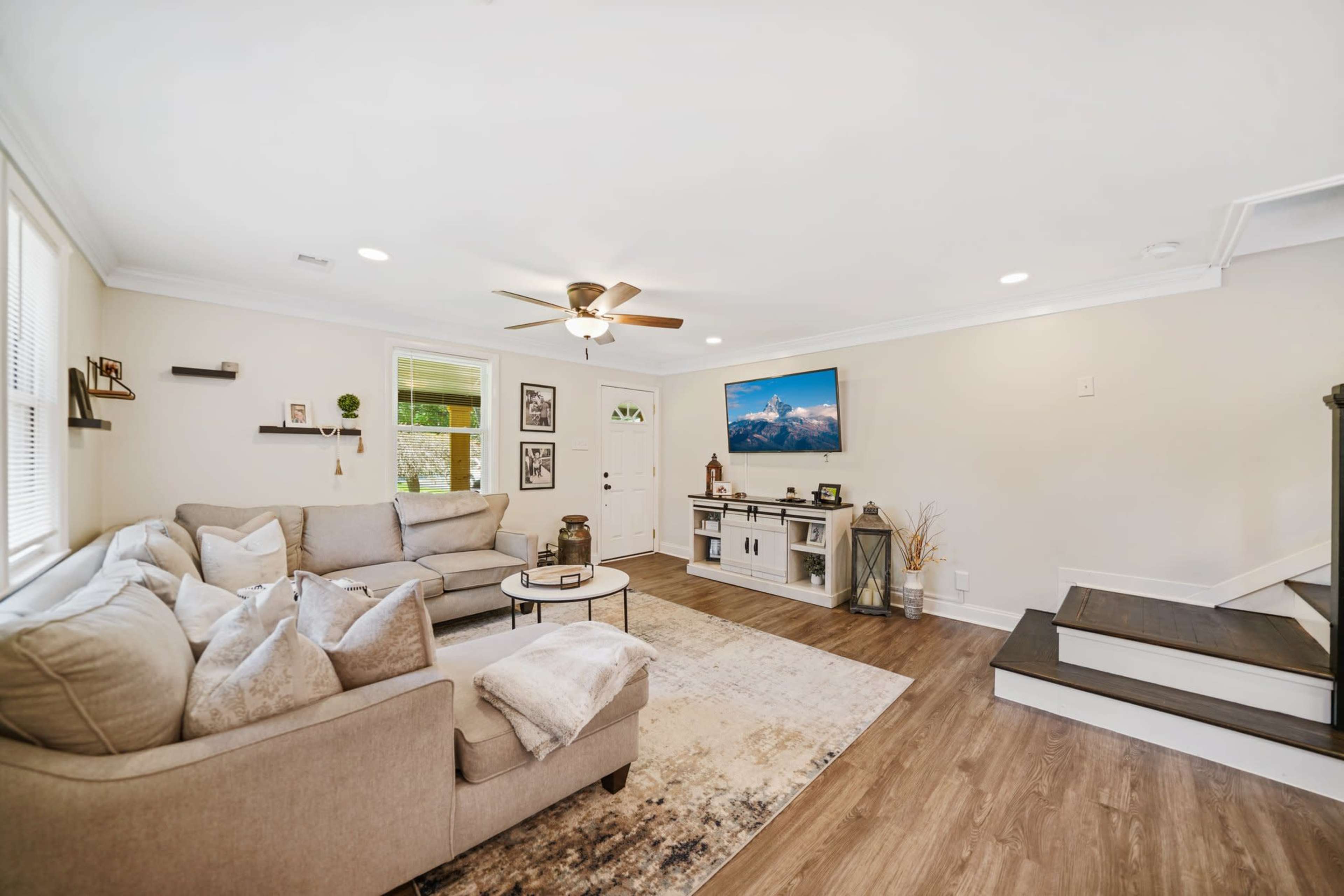 A living room features a gray sectional sofa, a round coffee table, a mounted television, and hardwood flooring, with a staircase visible in the background.