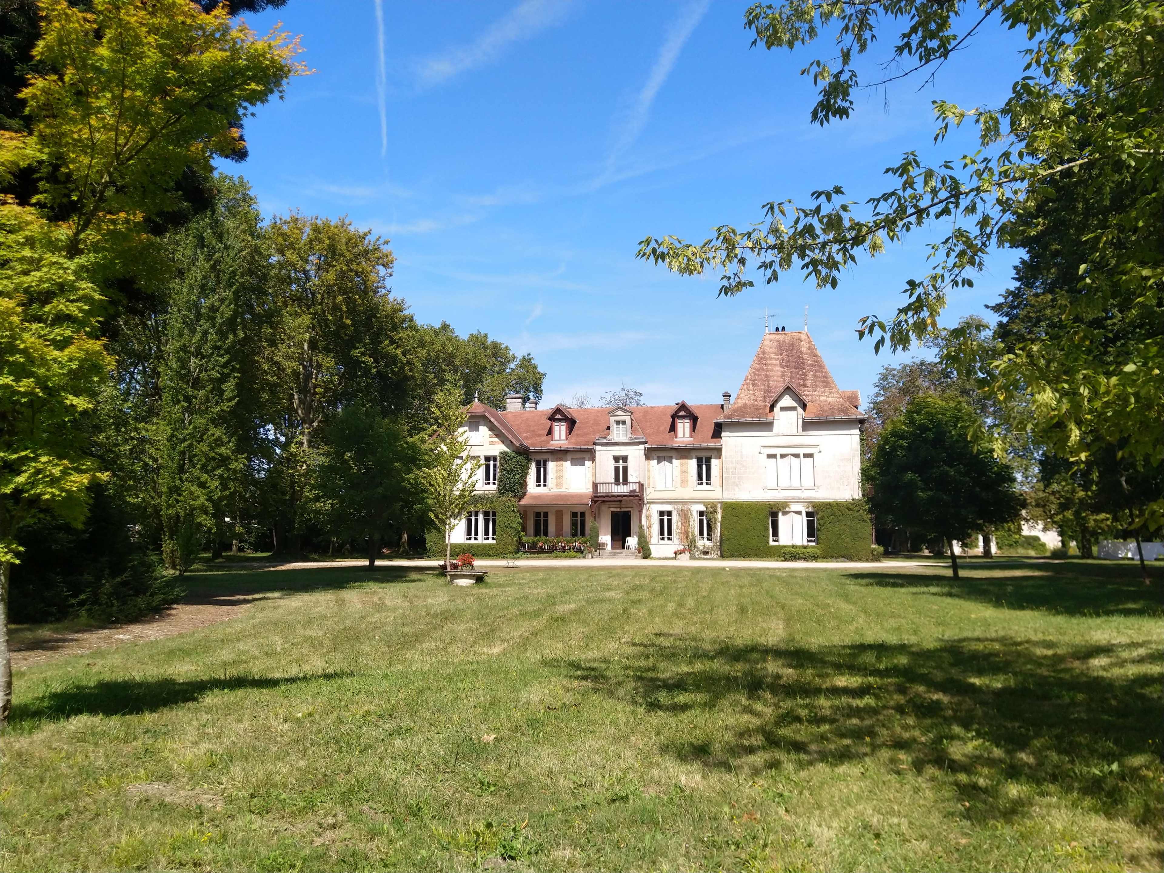 A large, two-story mansion with a red-tiled roof is set behind a grassy field surrounded by trees.