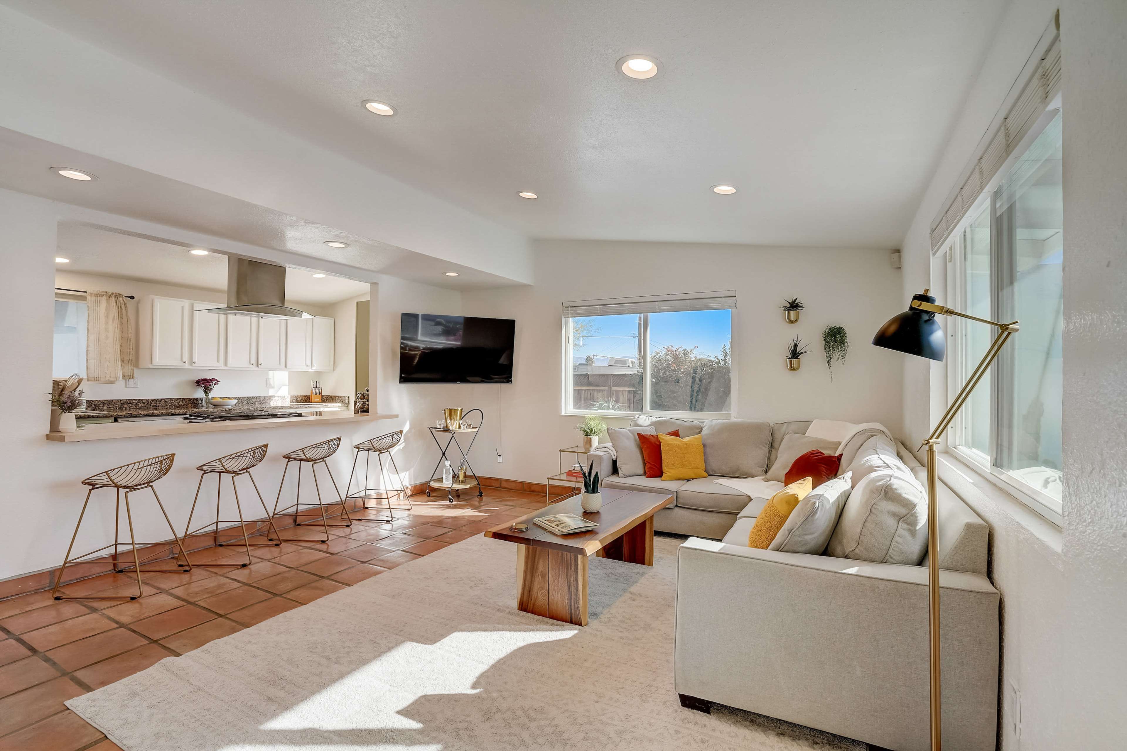 The living area features a light-colored sectional sofa, a wooden coffee table, and a kitchen bar with four metal stools, all arranged around large windows letting in natural light.