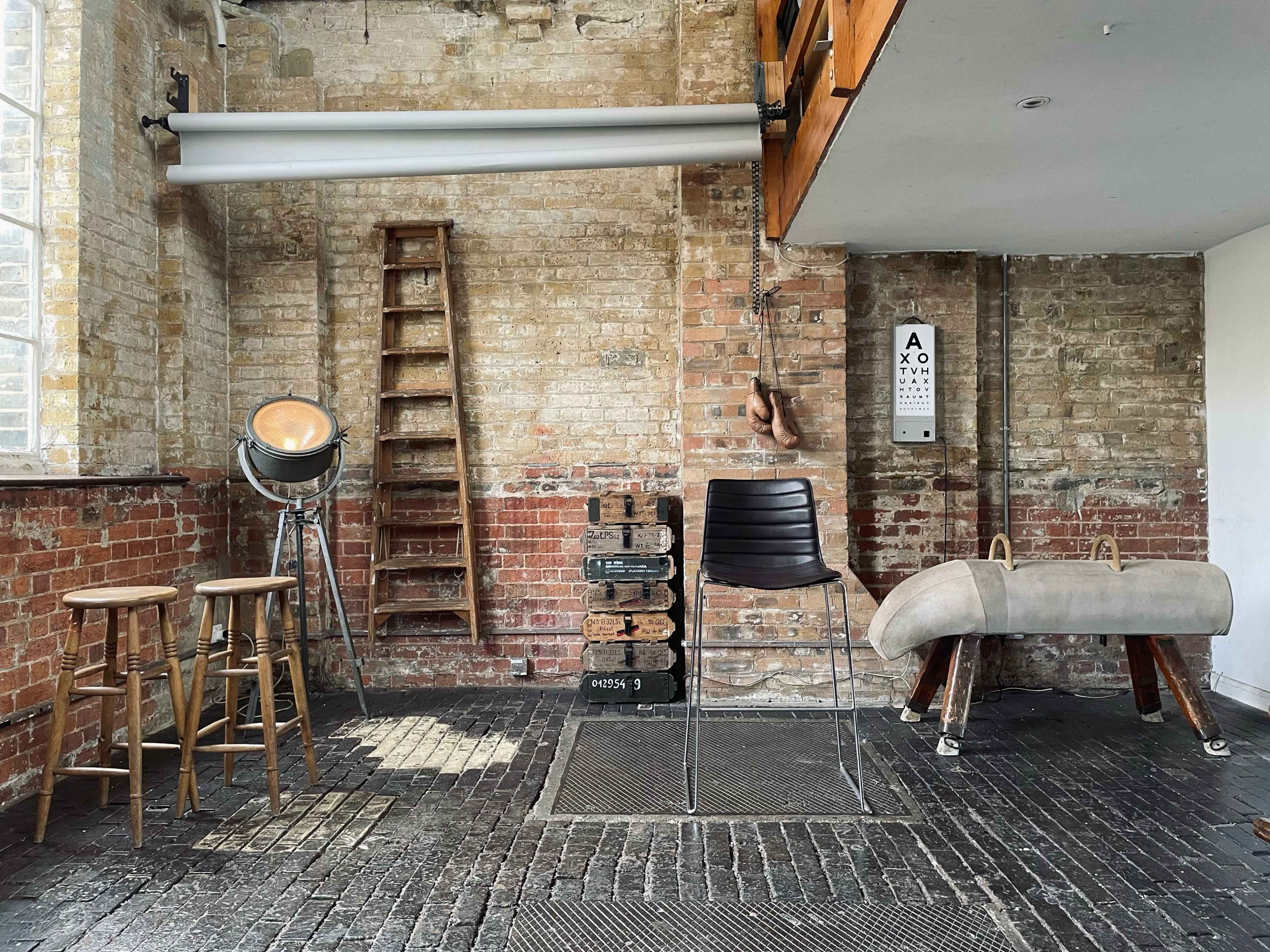 An industrial-style room with exposed brick walls, wooden ladders, a spotlight, a black chair, two wooden stools, and a vintage pommel horse.
