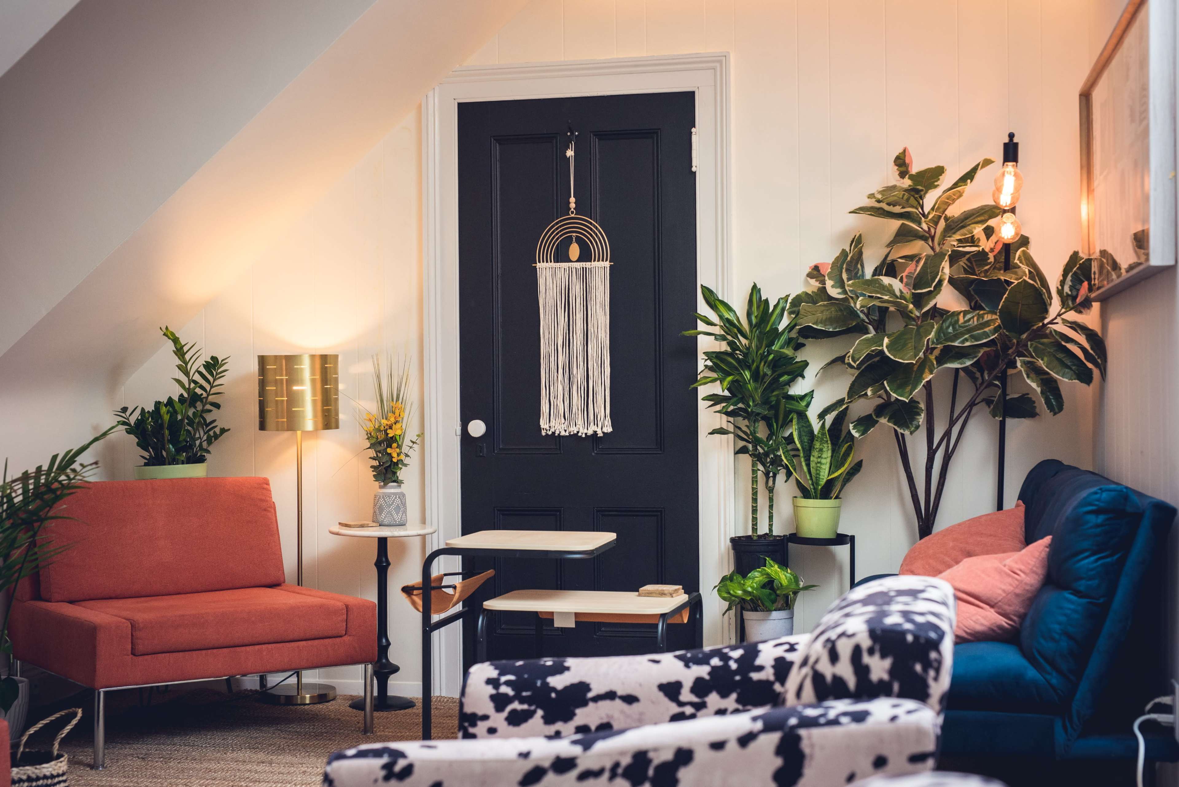 A cozy living room with two armchairs and a small side table, surrounded by indoor plants and featuring a black door with a hanging décor piece.