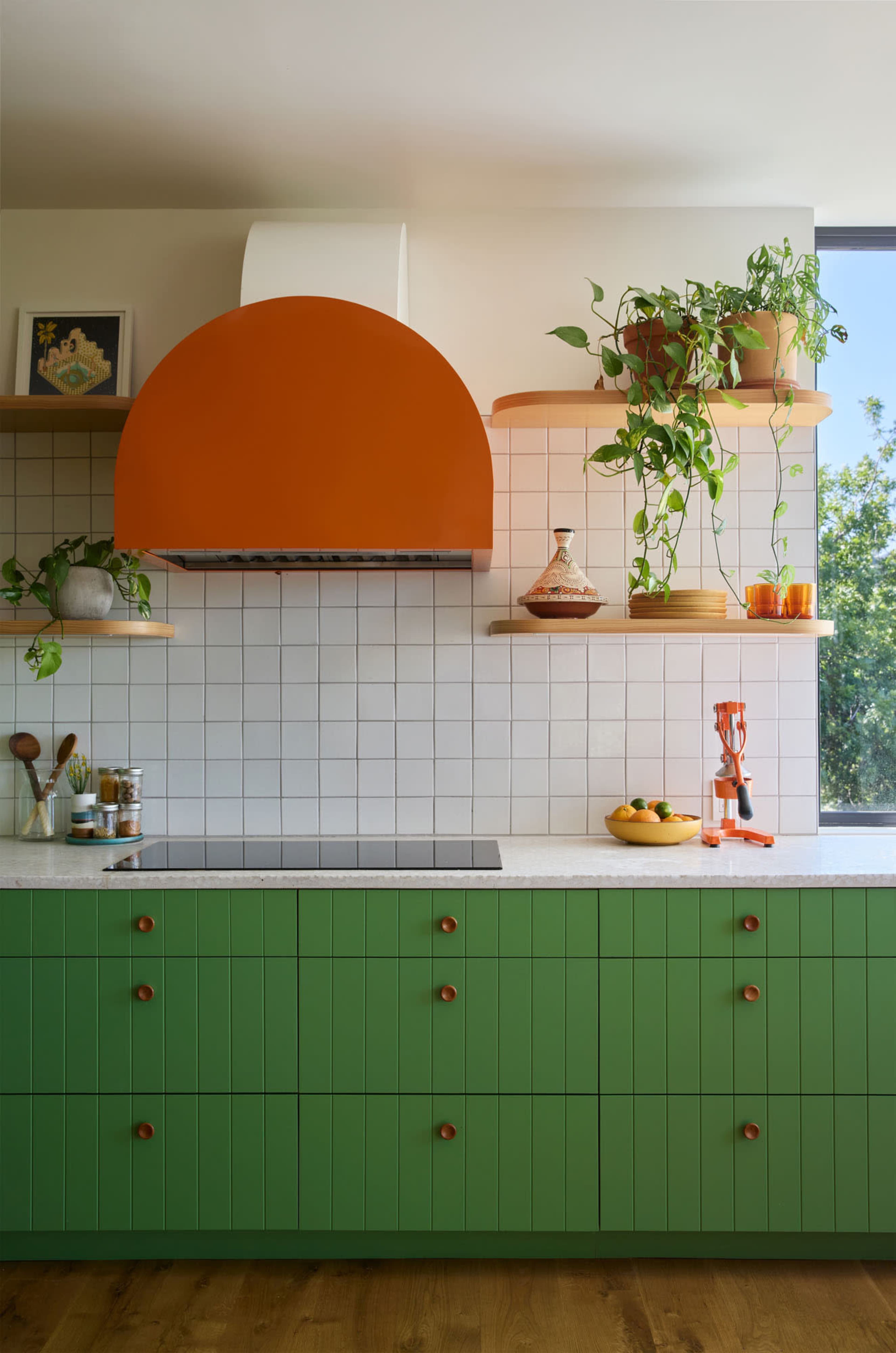 The image features a modern kitchen with green cabinetry, an orange hood above the stove, and open shelves displaying plants and kitchen items.