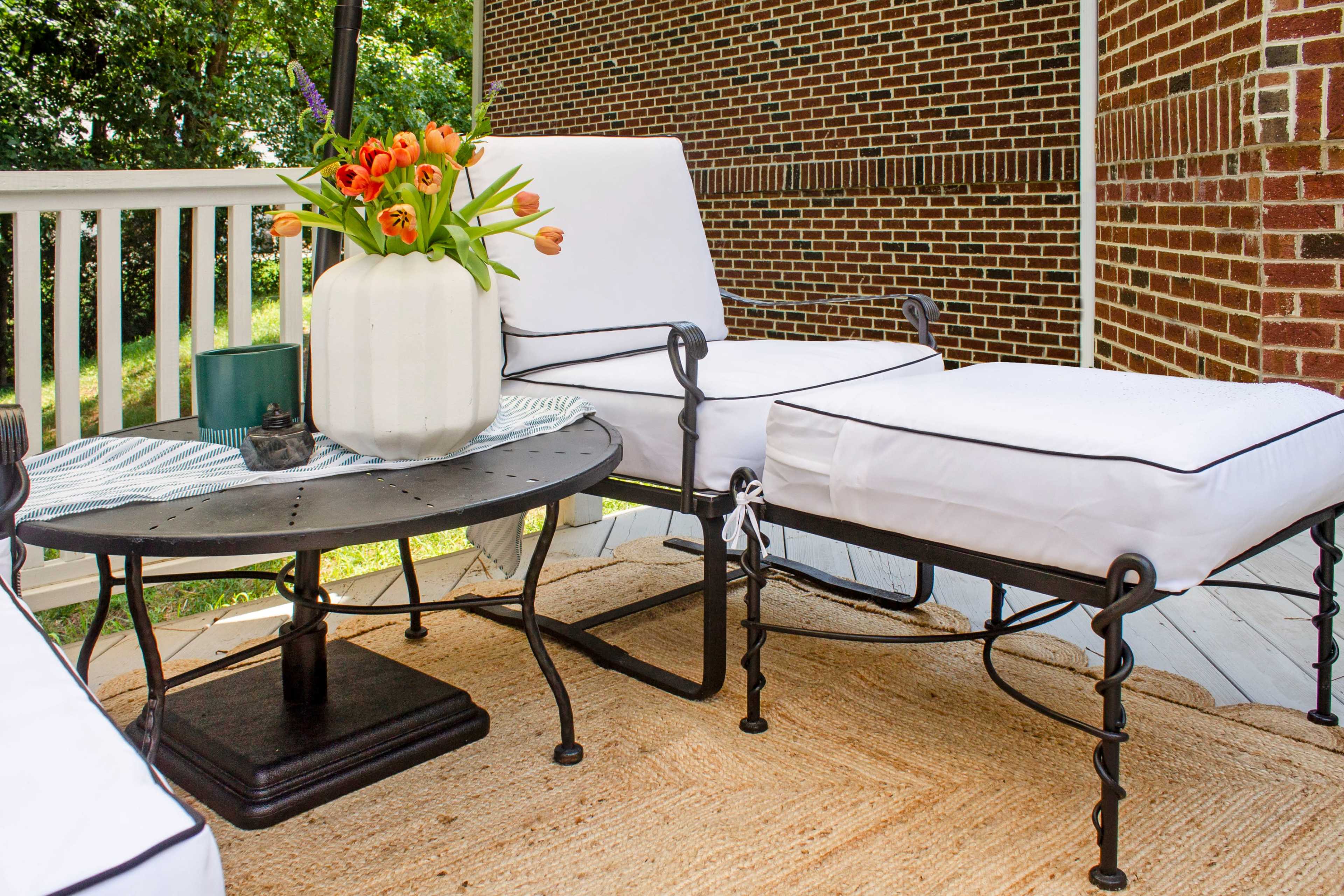 The image shows a patio setup featuring two white cushioned lounge chairs, a round black table, and a vase of orange tulips on top, all situated on a deck with a woven rug and a brick wall backdrop.
