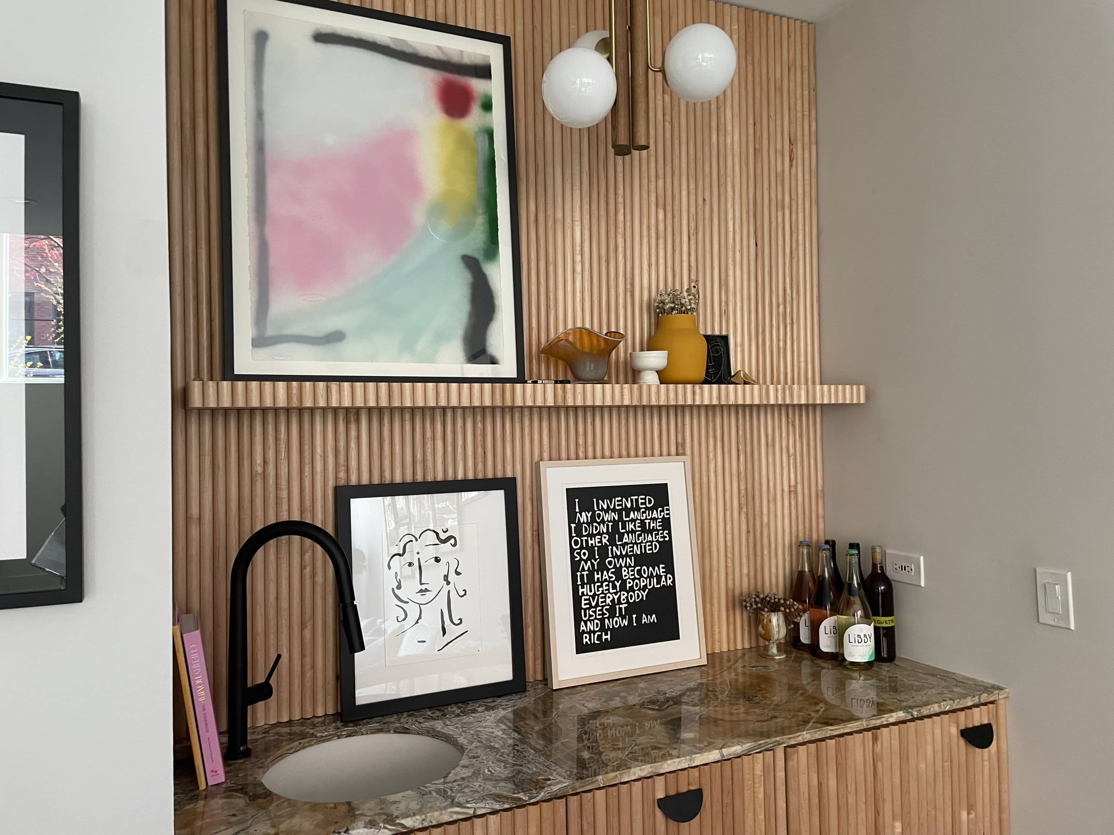 The image shows a modern bar area featuring wooden paneling, a granite countertop, framed artwork, and several bottles arranged on the shelves.