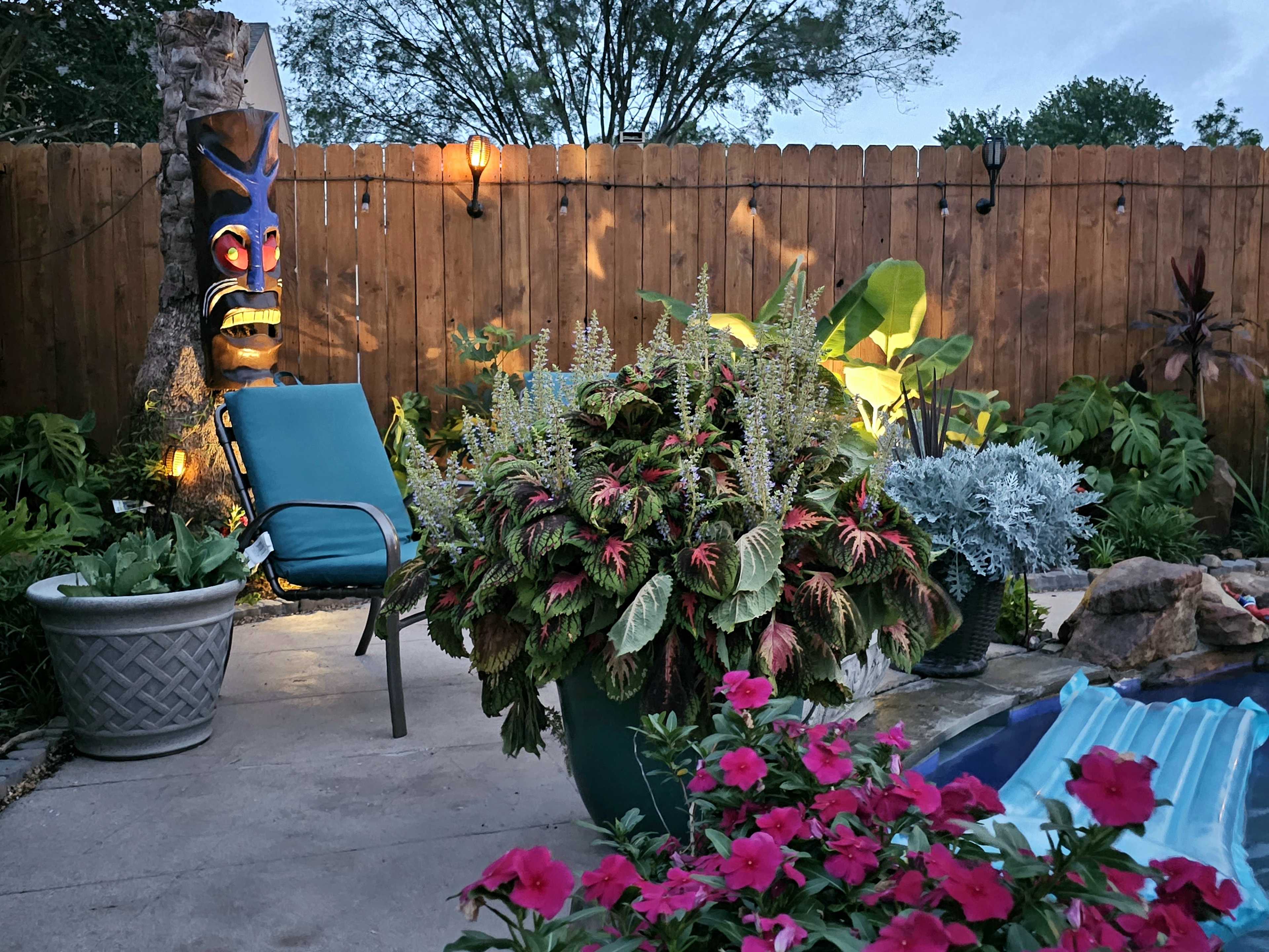 A garden area with a tiki statue, a lounge chair, various potted plants, and flowers near a pool surrounded by a wooden fence.