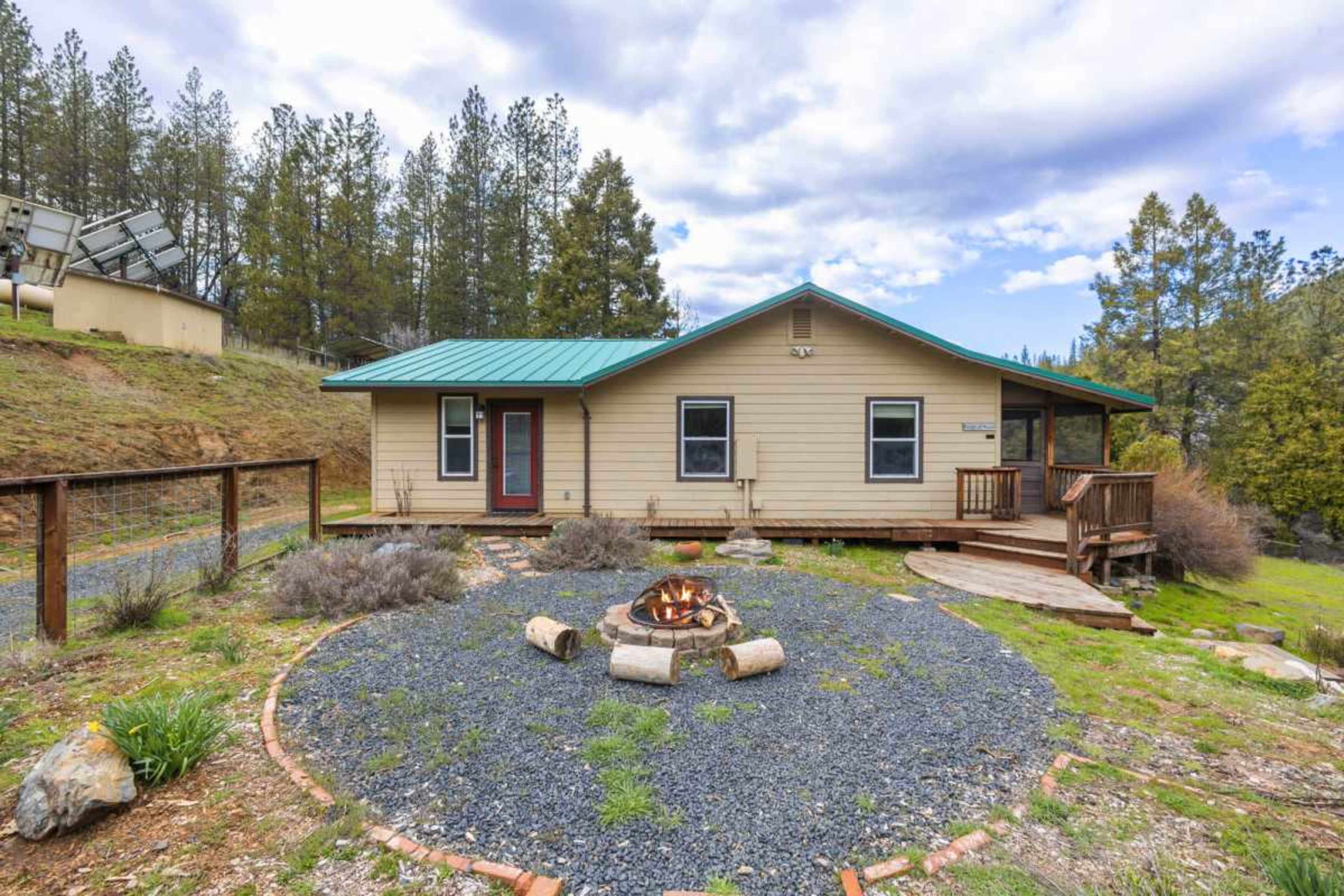 A single-story house with a teal metal roof is surrounded by gravel and a circular fire pit, set against a backdrop of pine trees.
