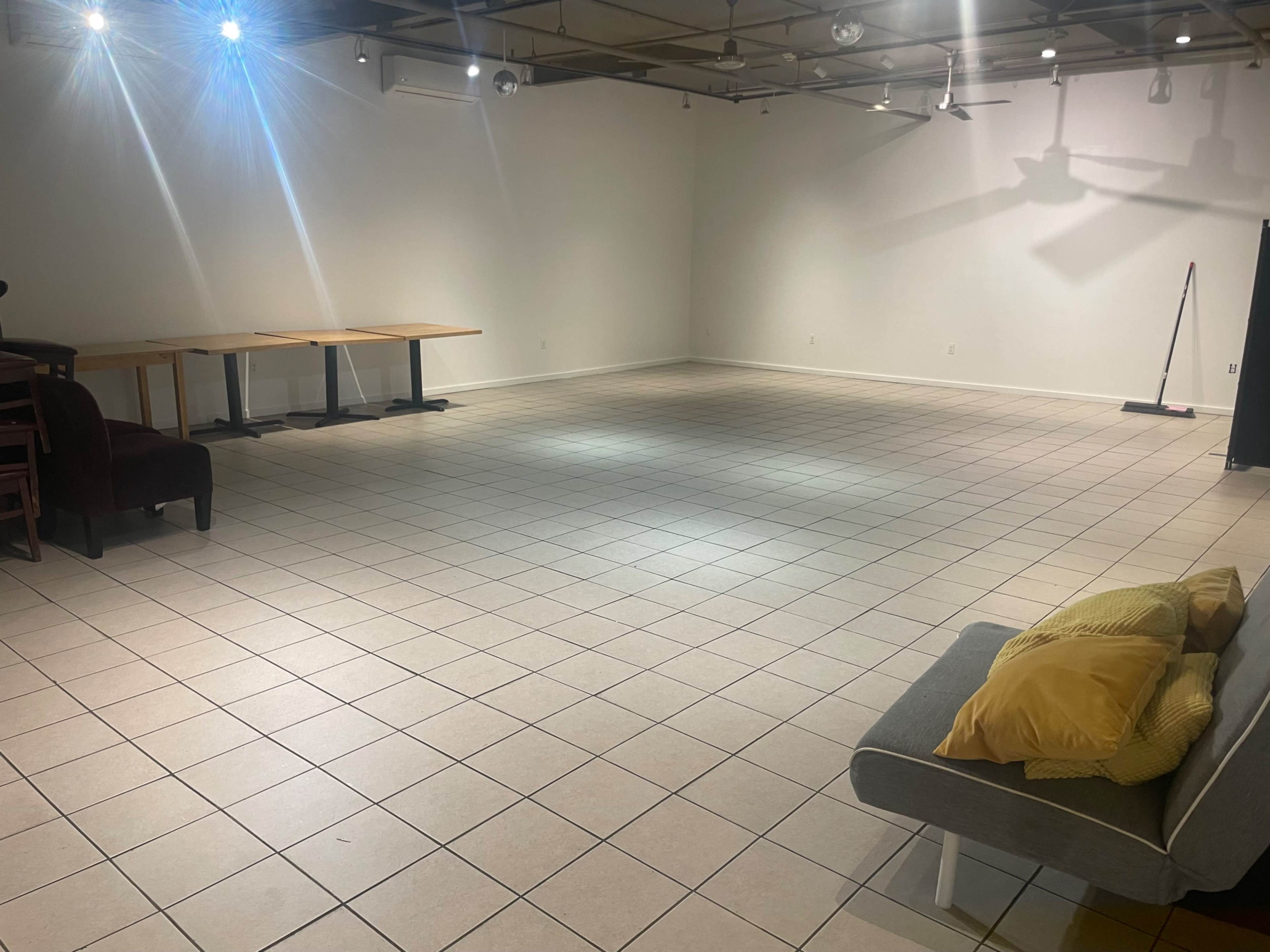 A spacious, empty room with tiled flooring, some tables lined against the wall, and a sofa with yellow cushions in one corner.