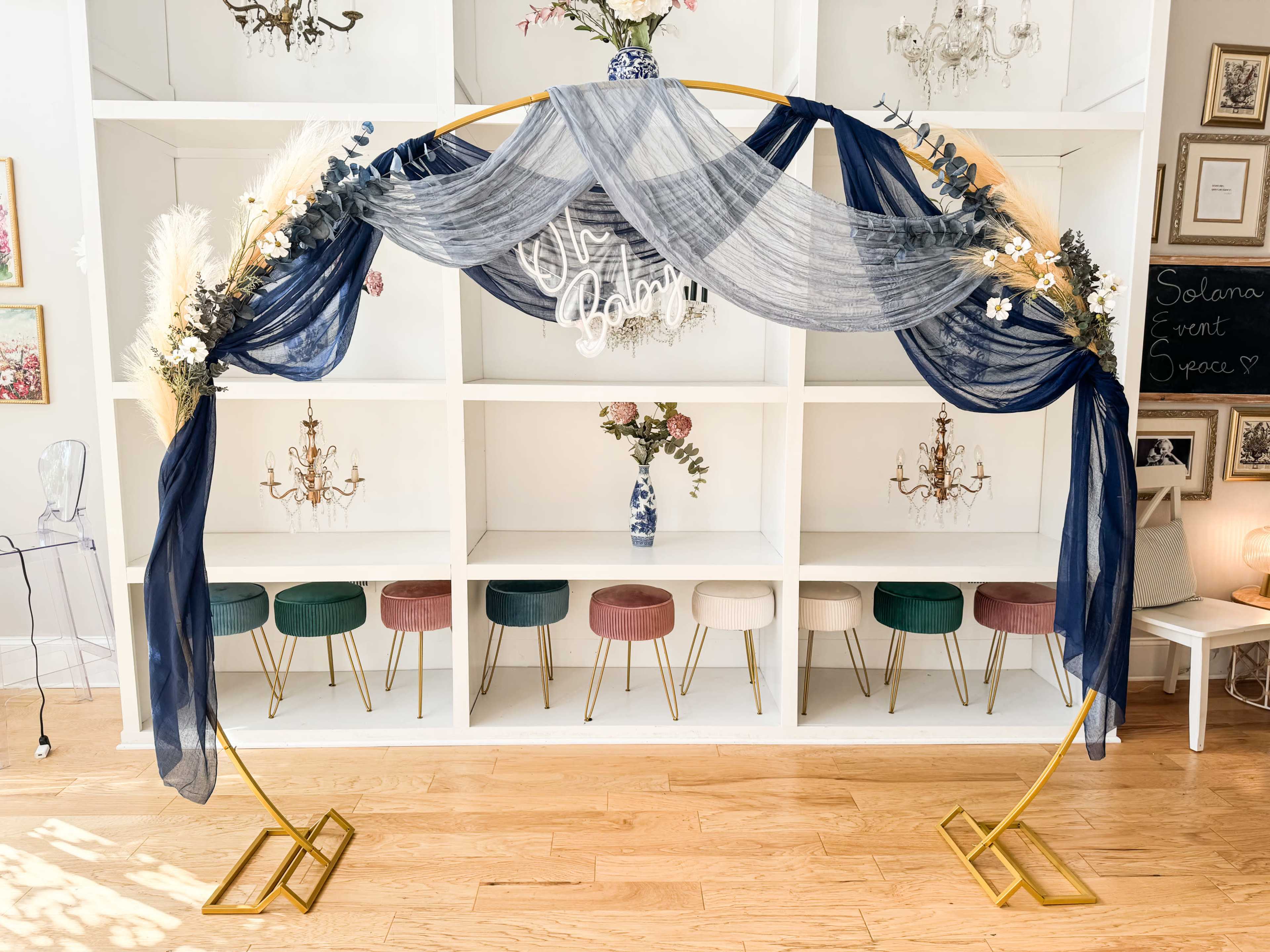 The image shows a decorative arch with navy fabric and floral arrangements in front of a white, shelf-like background featuring vases and colorful stools.