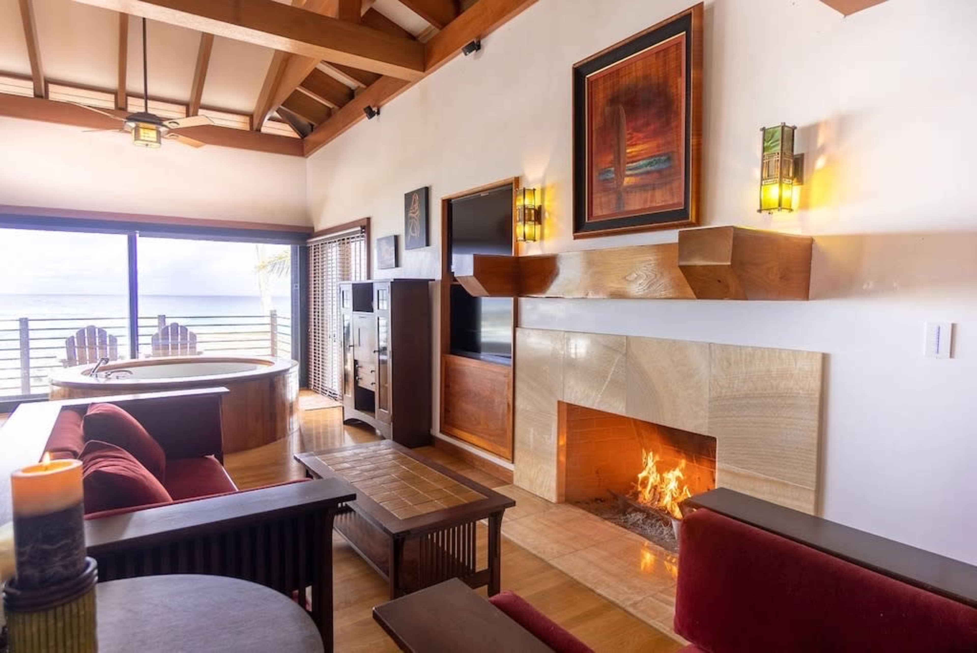 A cozy living room features a fireplace, wooden ceiling beams, and large windows overlooking a view of the ocean.