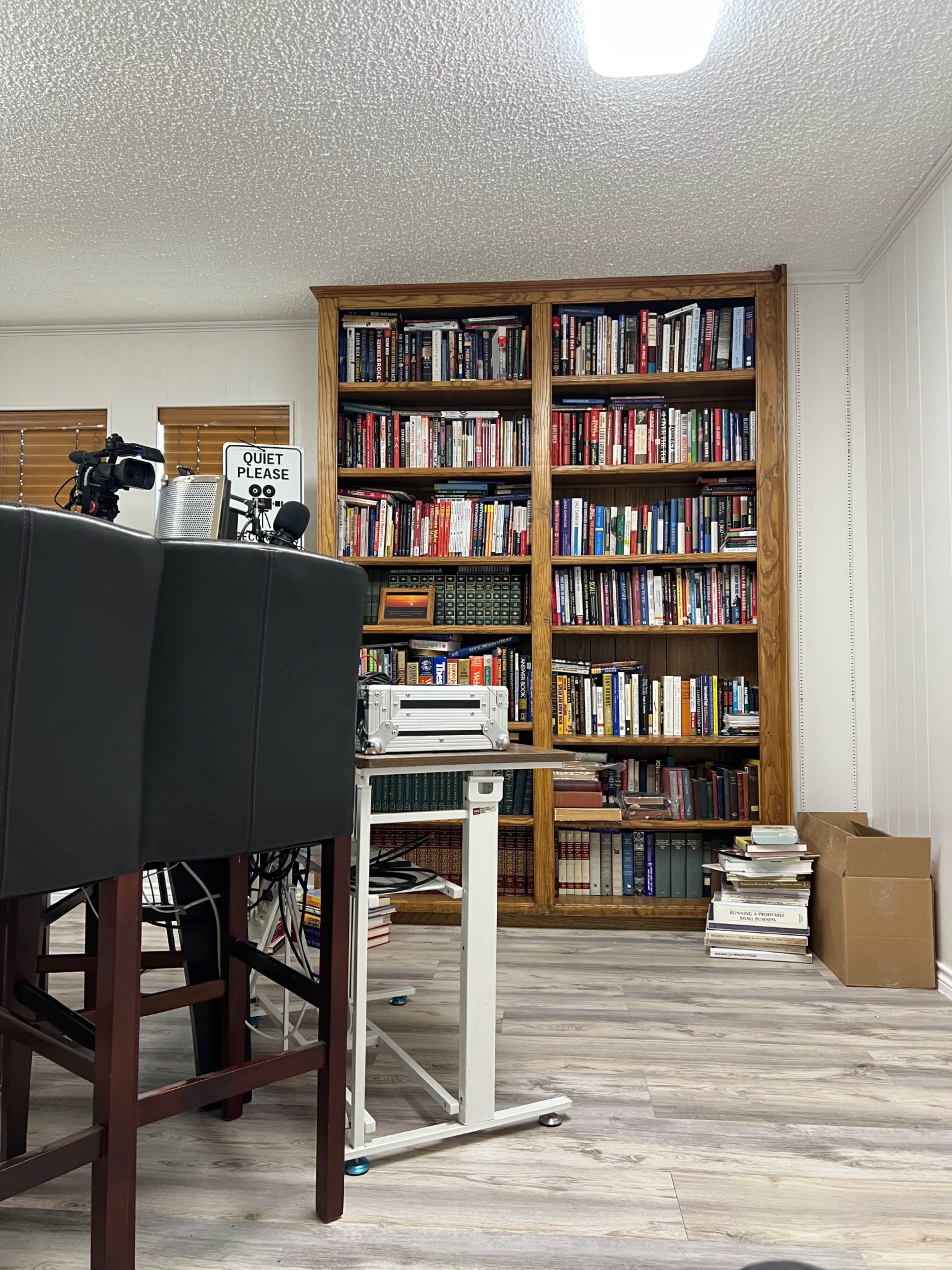 A large bookshelf filled with books stands against a wall in a room that features a table and rolling cart.