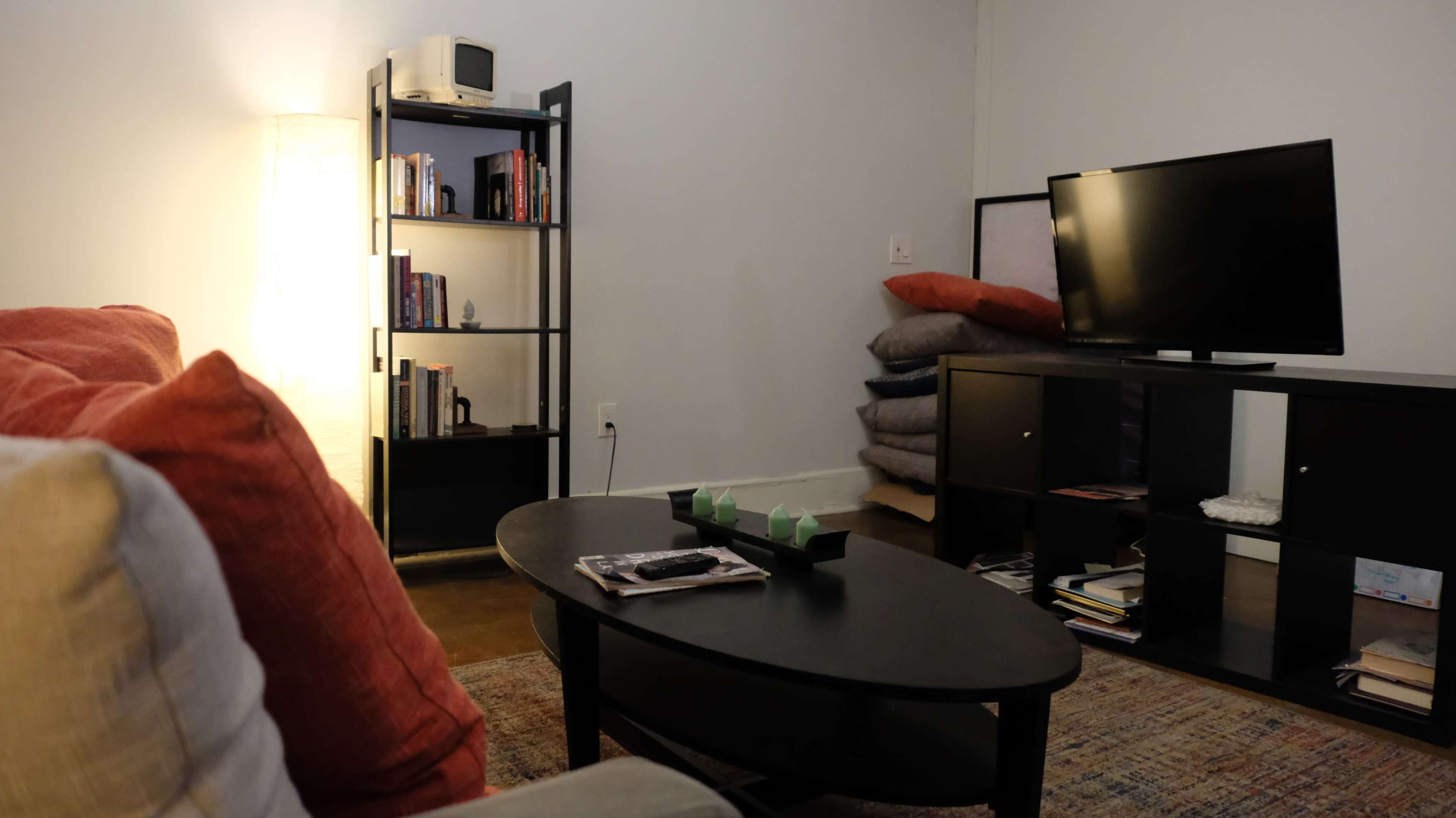 The image shows a cozy living room featuring a couch, a black coffee table, a bookshelf filled with books, and a television on a stand, with stacks of cushions beside it.