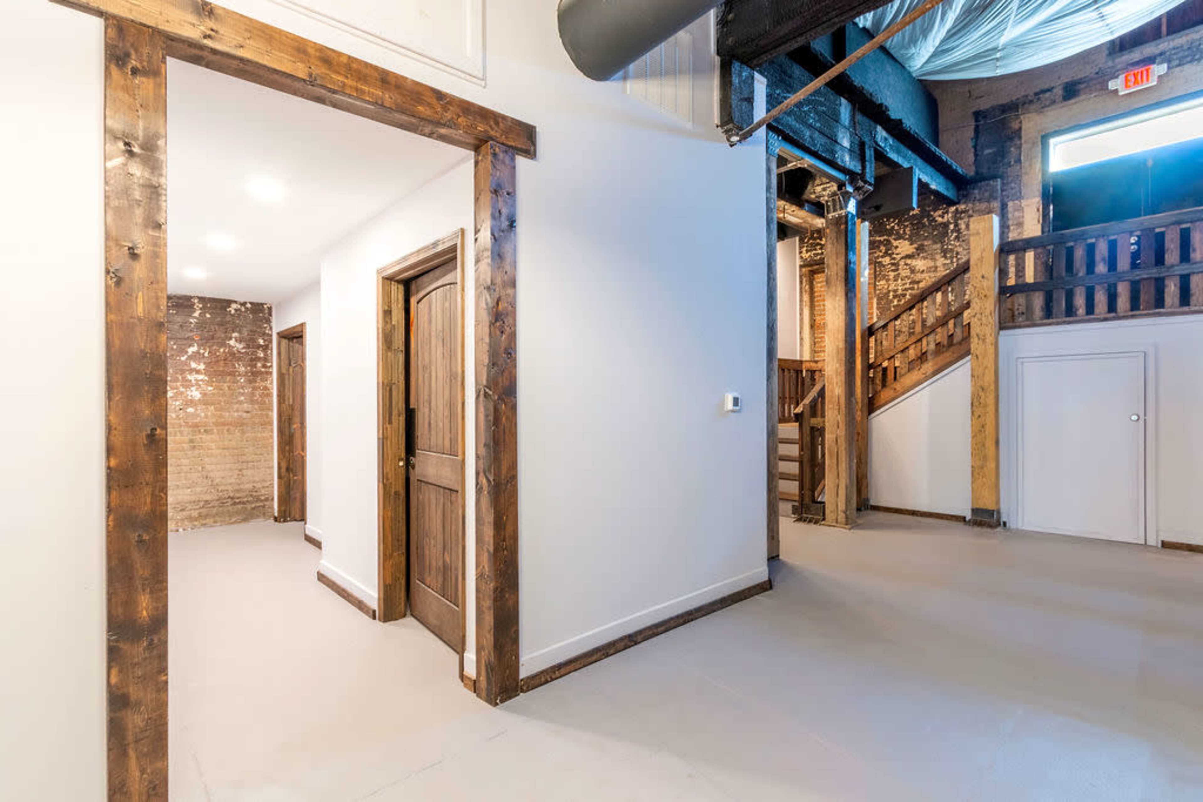 An interior space featuring a hallway leading to wooden doorways, with a staircase visible in the background and exposed brick walls.