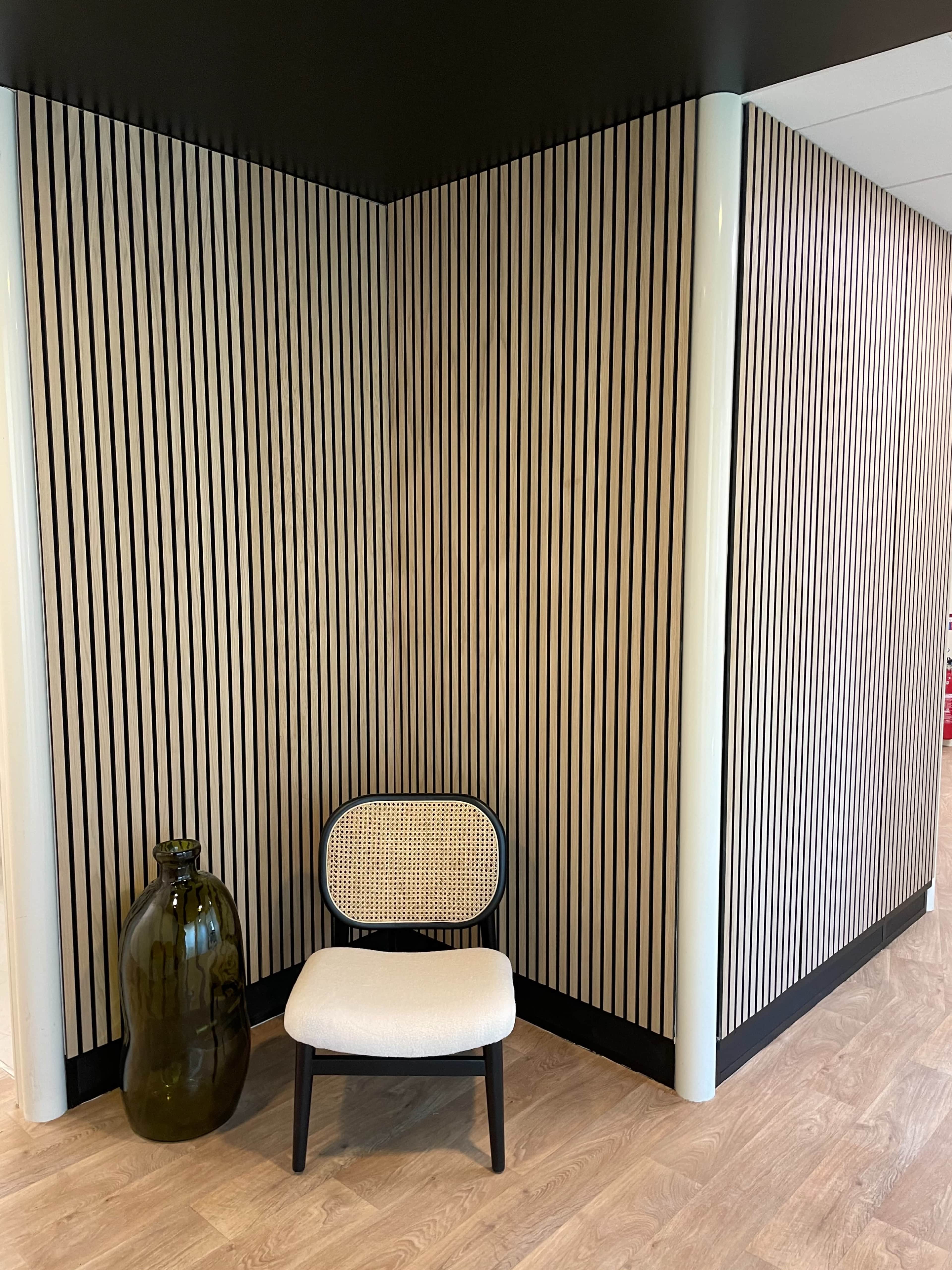 A corner of a room with vertical striped wood paneling, a round-backed chair with a cane seat, and a large green glass vase.