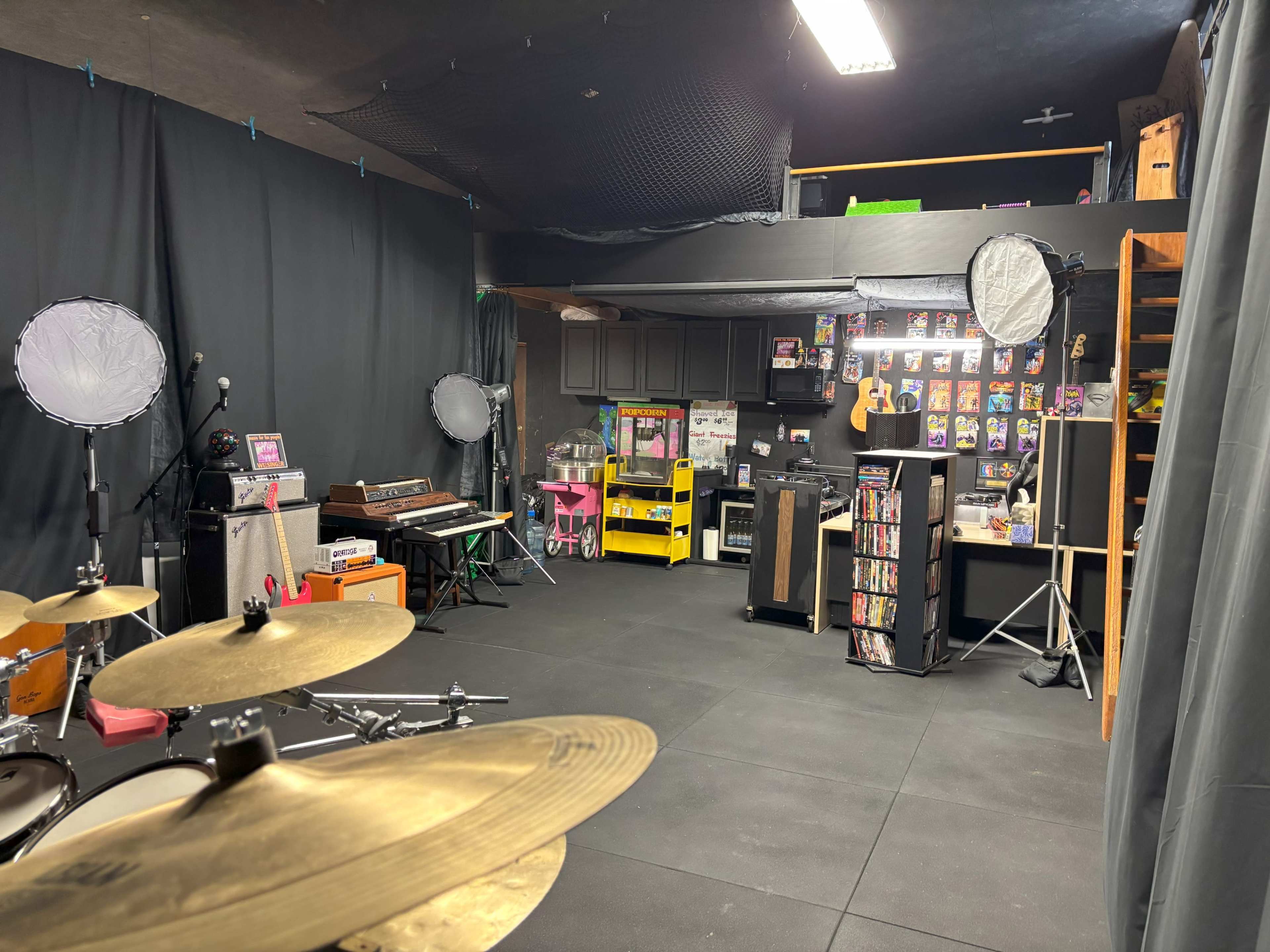 The image shows a music rehearsal space with various instruments, amplifiers, a keyboard, and studio lighting equipment against a backdrop of black curtains and decorated walls.