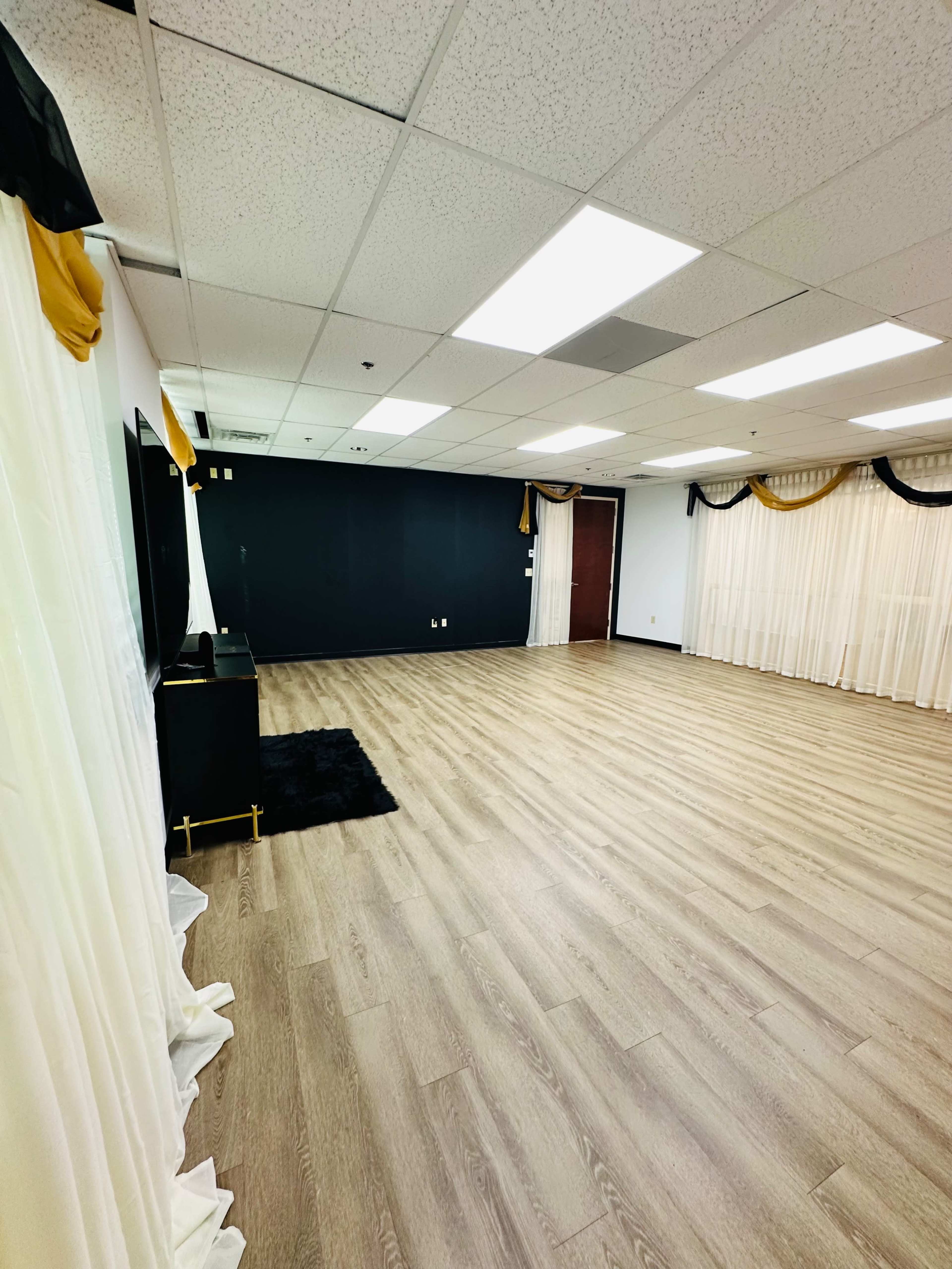 The image shows a spacious room with light wooden flooring, black and yellow decorations, and sheer curtains along one wall.