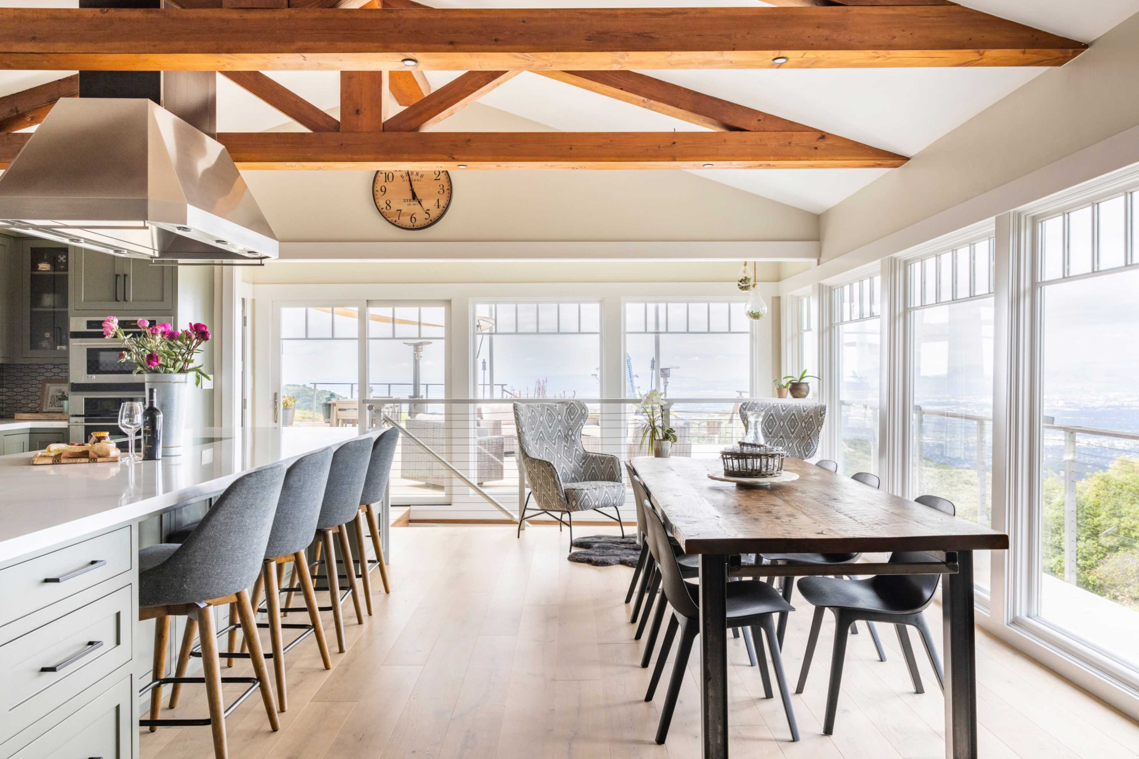 A modern kitchen features a long dining table and a bar with high stools, illuminated by large windows that offer a view of the outdoors.