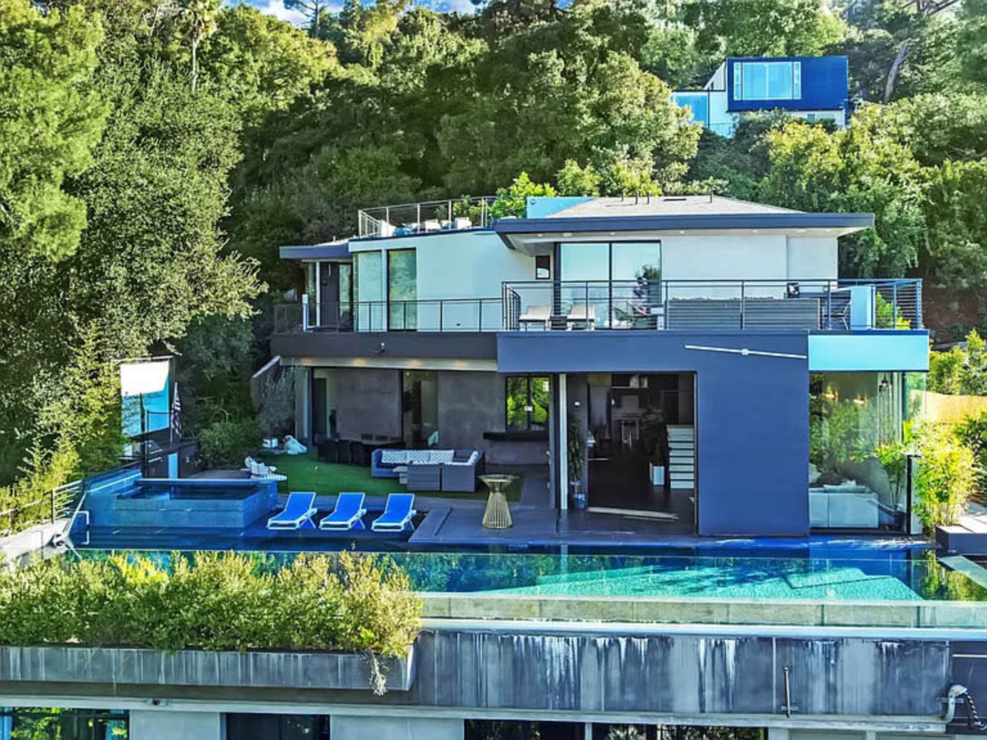 The image shows a modern house with large glass windows, a swimming pool, and outdoor seating, surrounded by lush greenery.