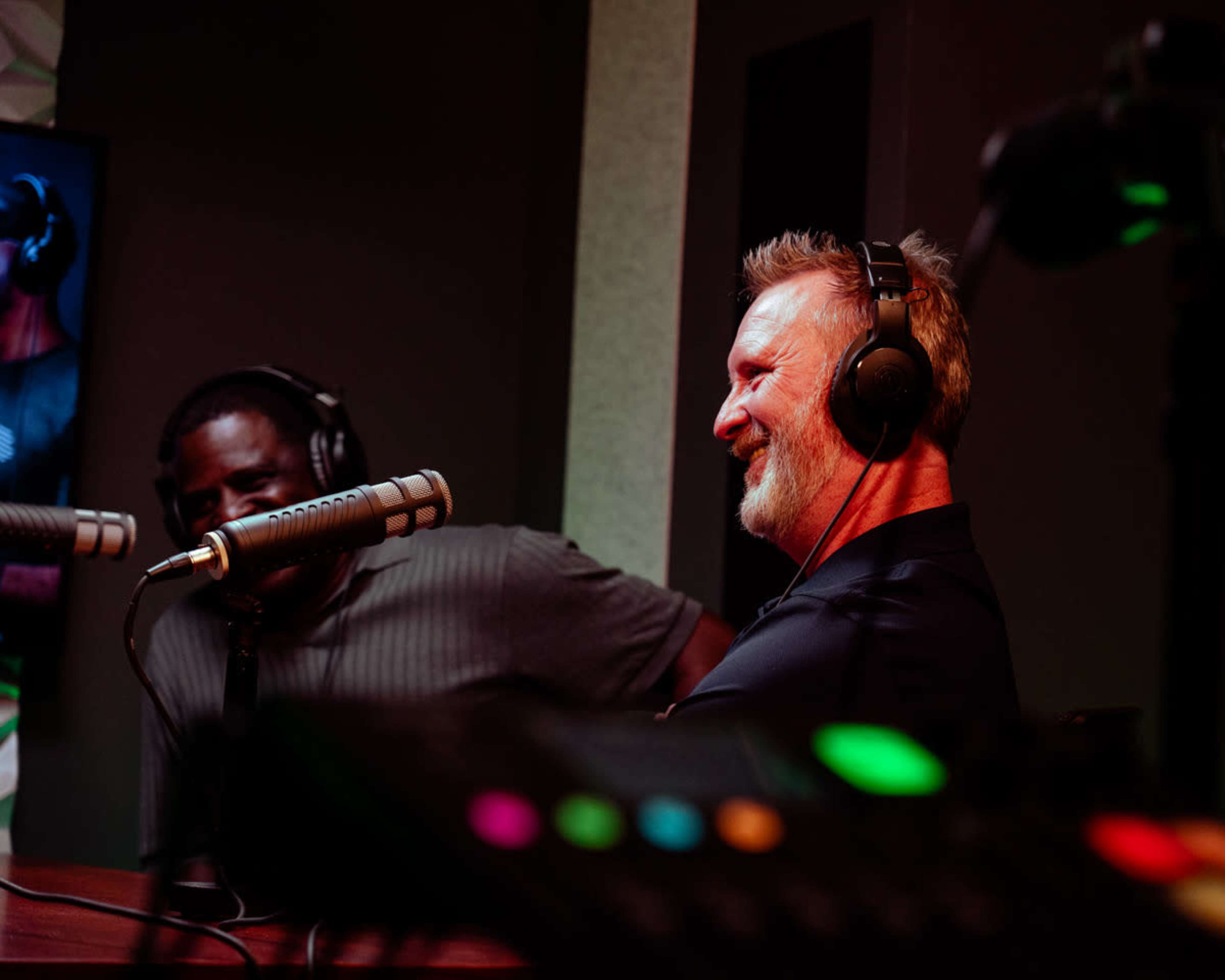 Two men wearing headphones engage in conversation at a podcast recording studio, with microphones and a control panel visible in the foreground.