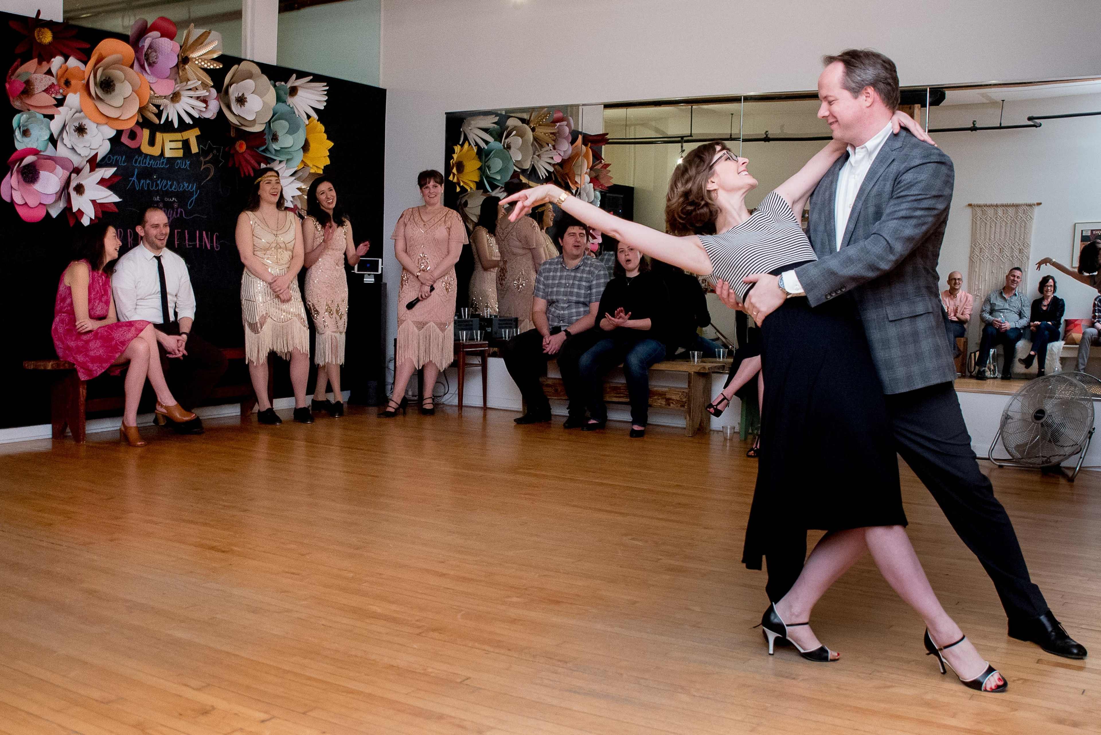 A couple performs a dance in front of an audience at a social event, while others sit and watch in the background.