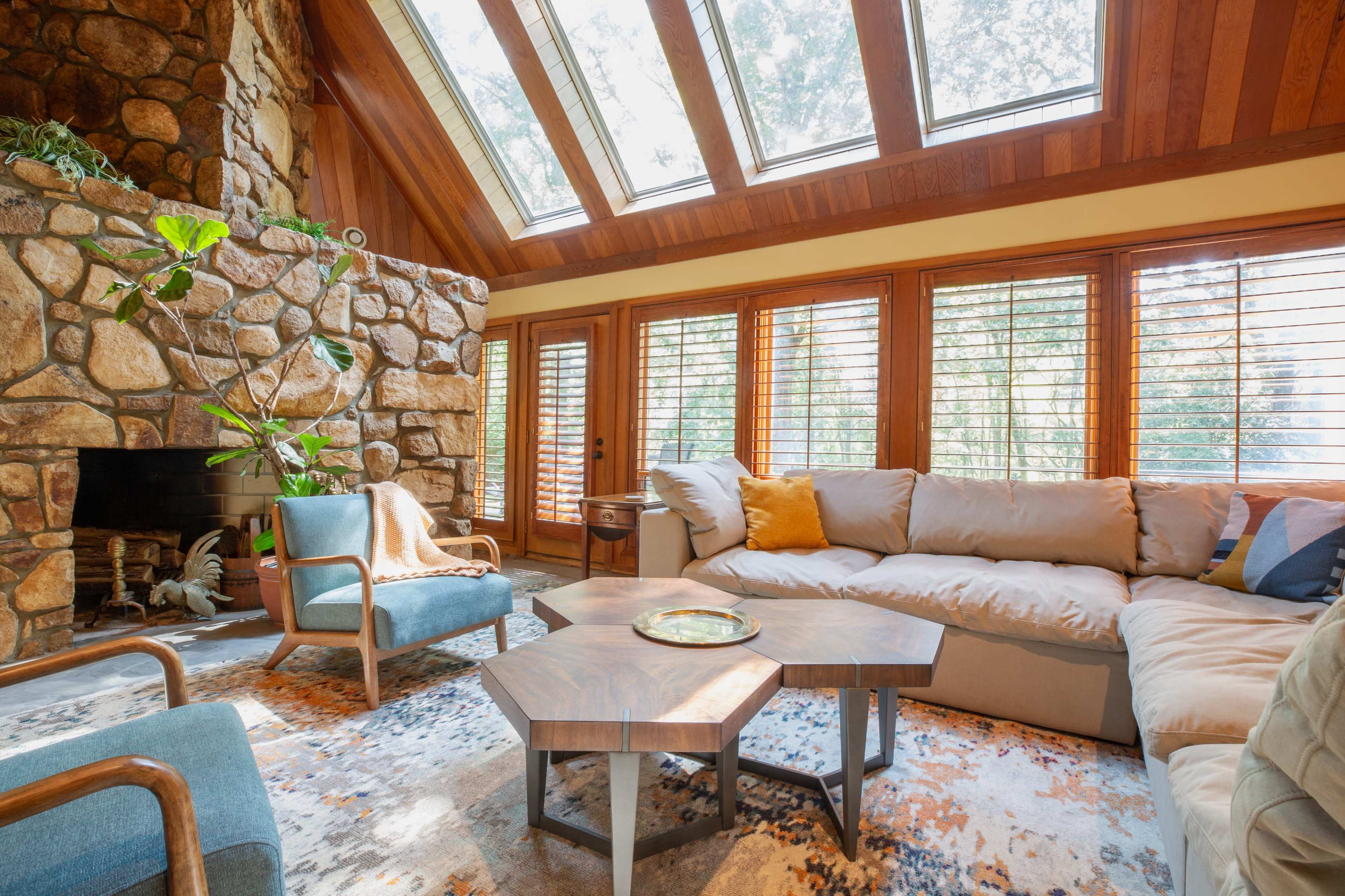 A spacious living room featuring a stone fireplace, large skylights, and a combination of modern furniture and a patterned area rug.
