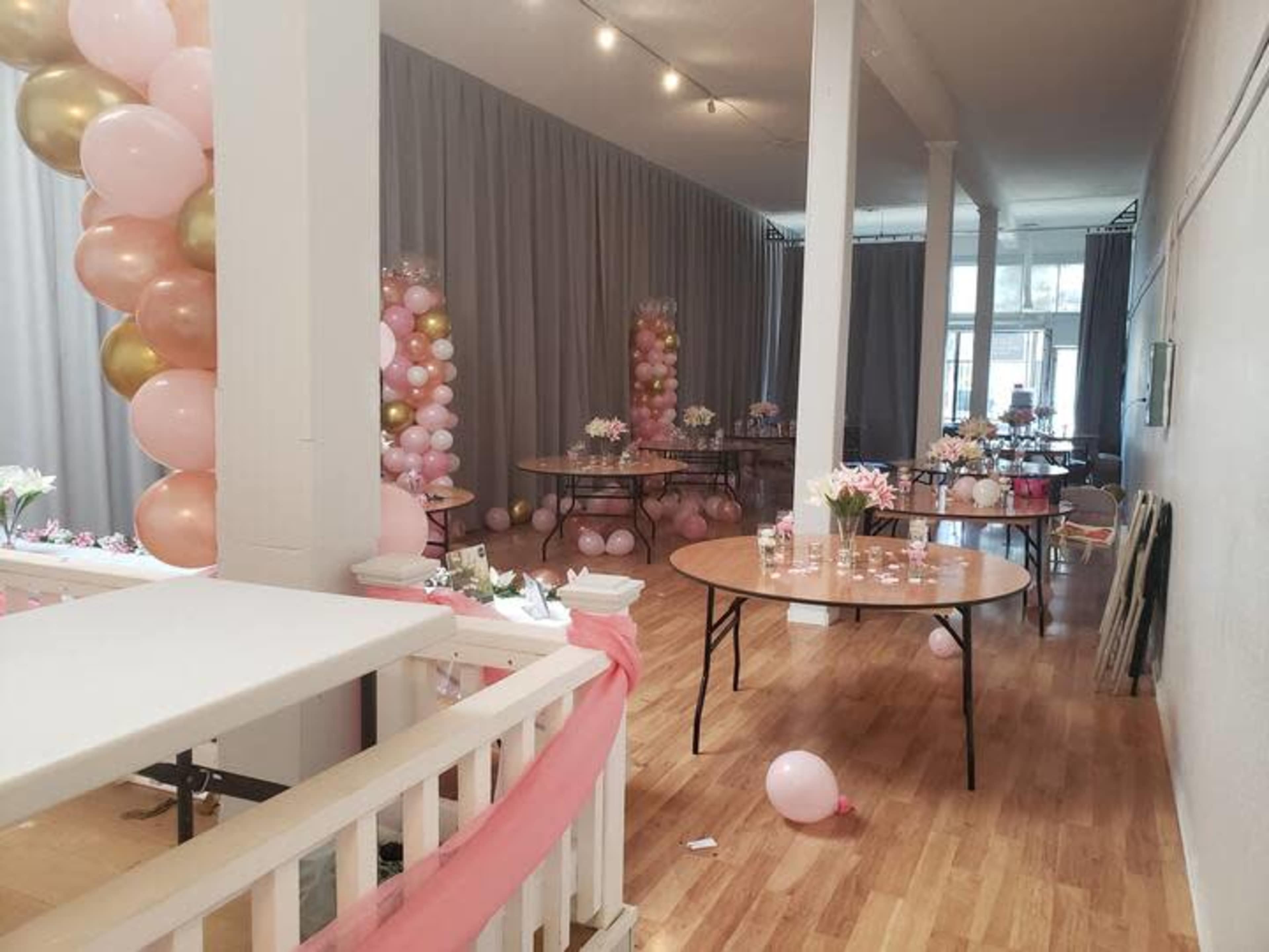 A decorated event space with round tables, floral centerpieces, and balloon arrangements in pink and gold.