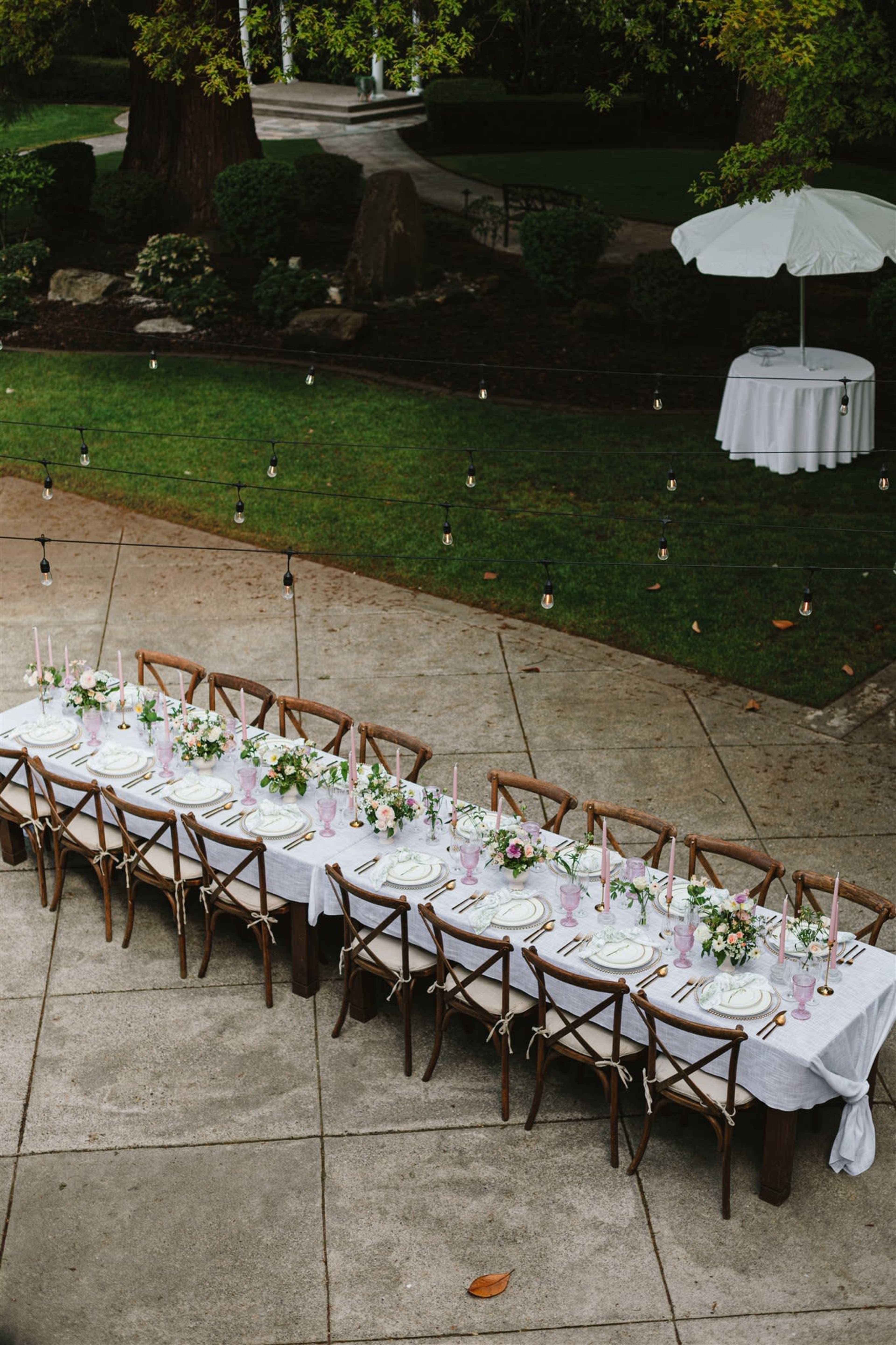 A long, elegantly set dining table with floral arrangements and candles is arranged outdoors on a stone patio, surrounded by greenery.