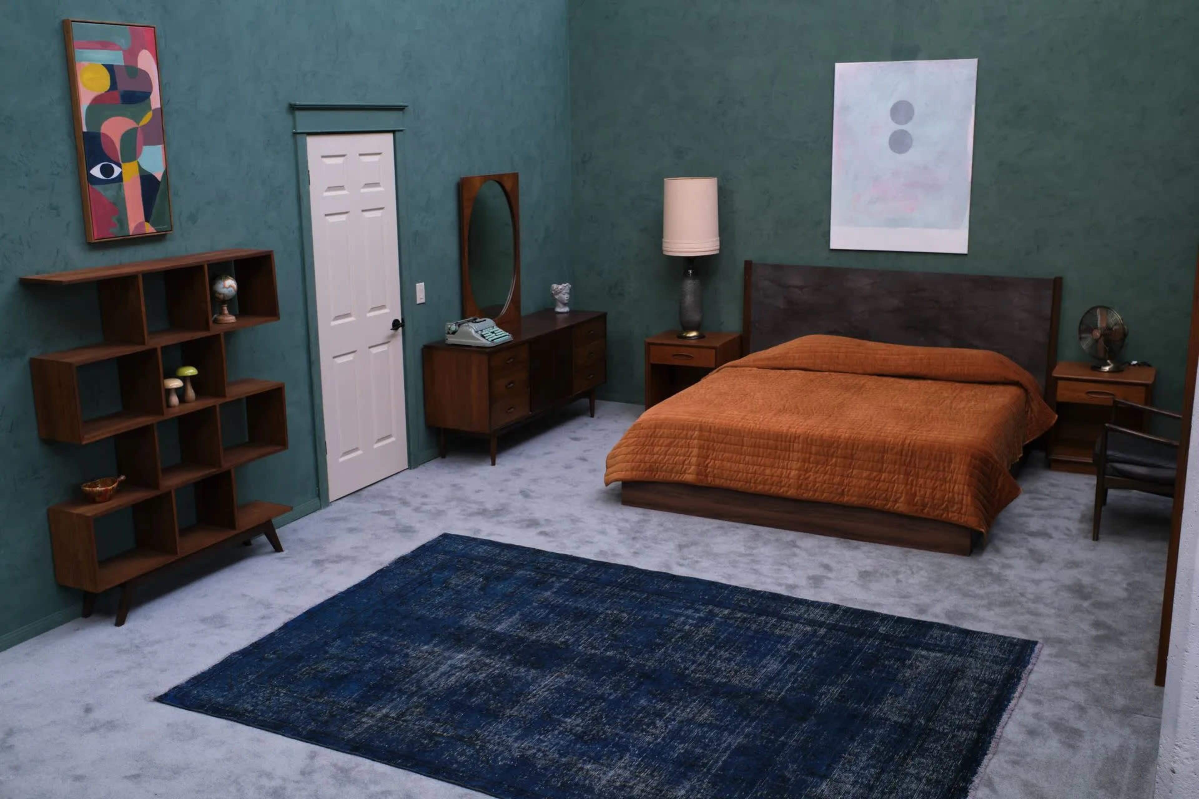 The scene depicts a modern bedroom with a large bed, wooden furniture, a rug, and a muted green wall.