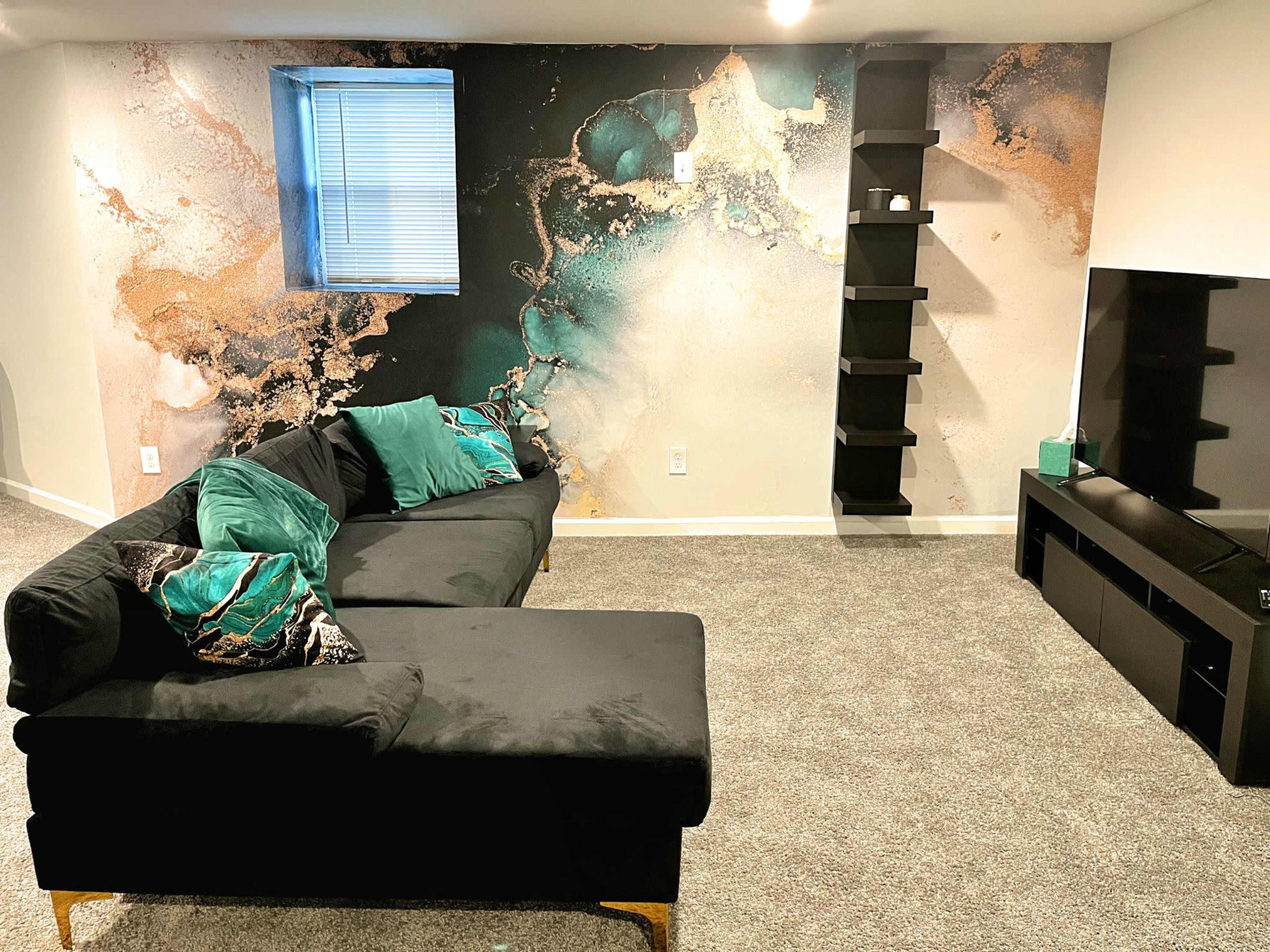 A cozy living room features a black sectional sofa, a textured wall mural with abstract designs, and a black TV stand against a light-colored wall.