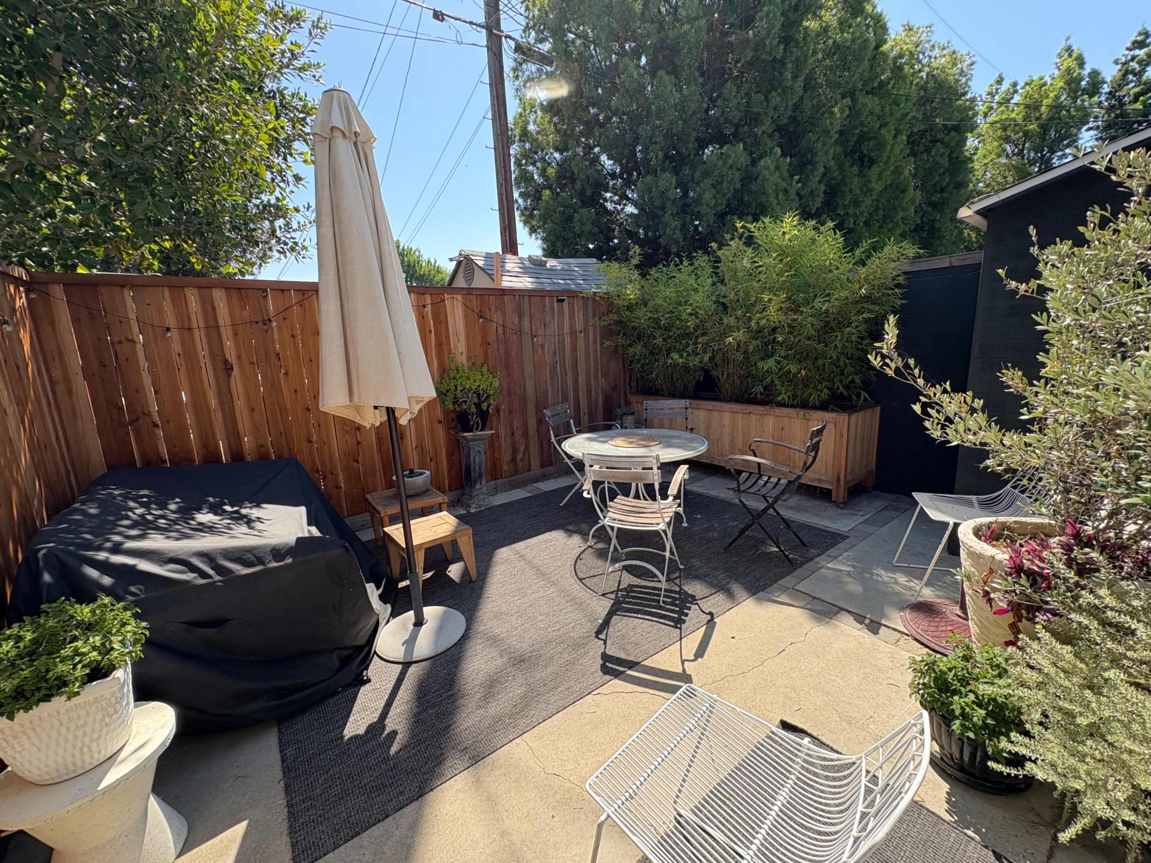 The image shows a small outdoor patio area furnished with a round table and chairs, a large umbrella, and various potted plants, surrounded by wooden fencing and vegetation.