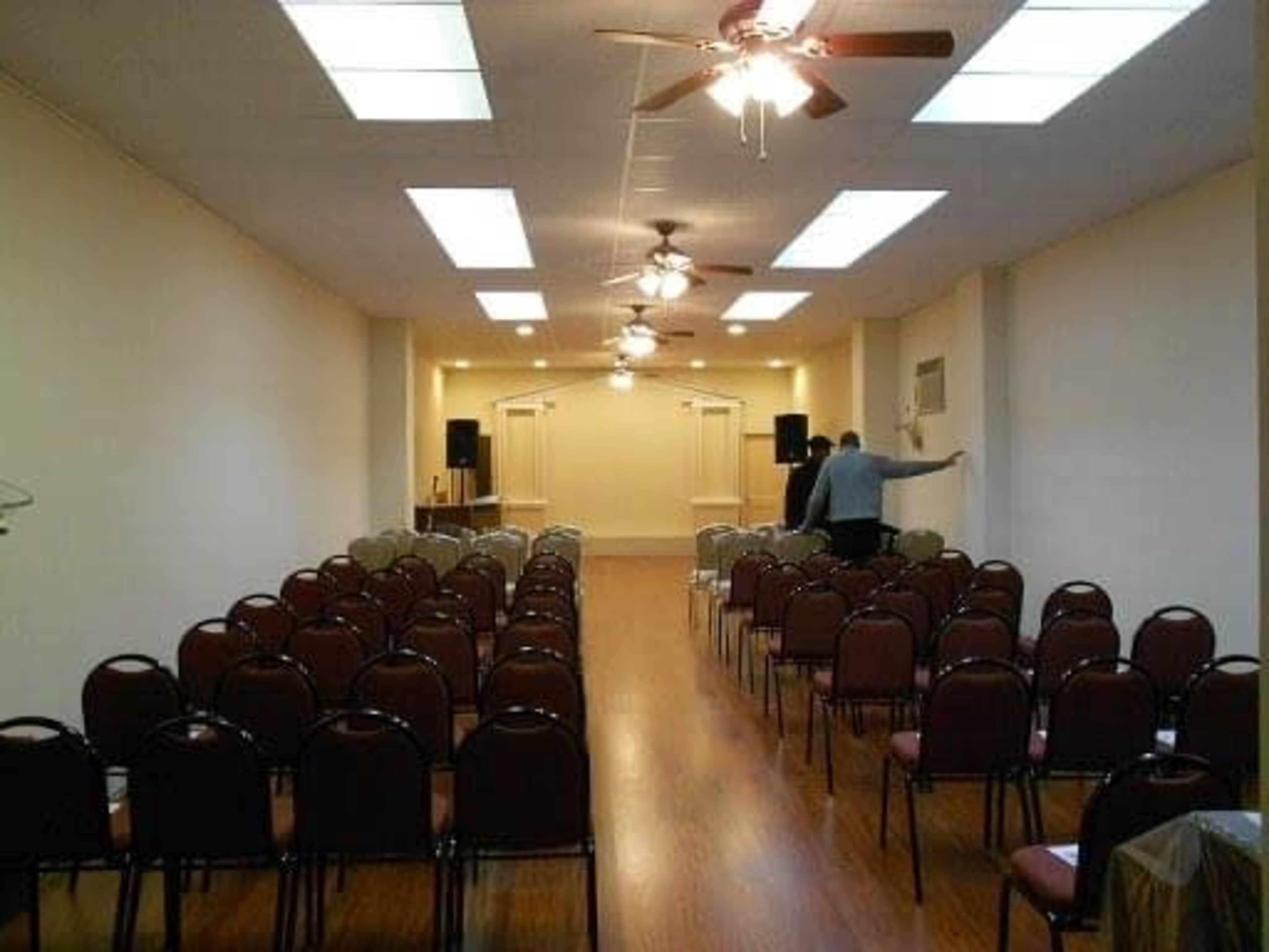 A large, empty room set up with rows of chairs facing a stage area, with ceiling fans and speakers positioned along the walls.
