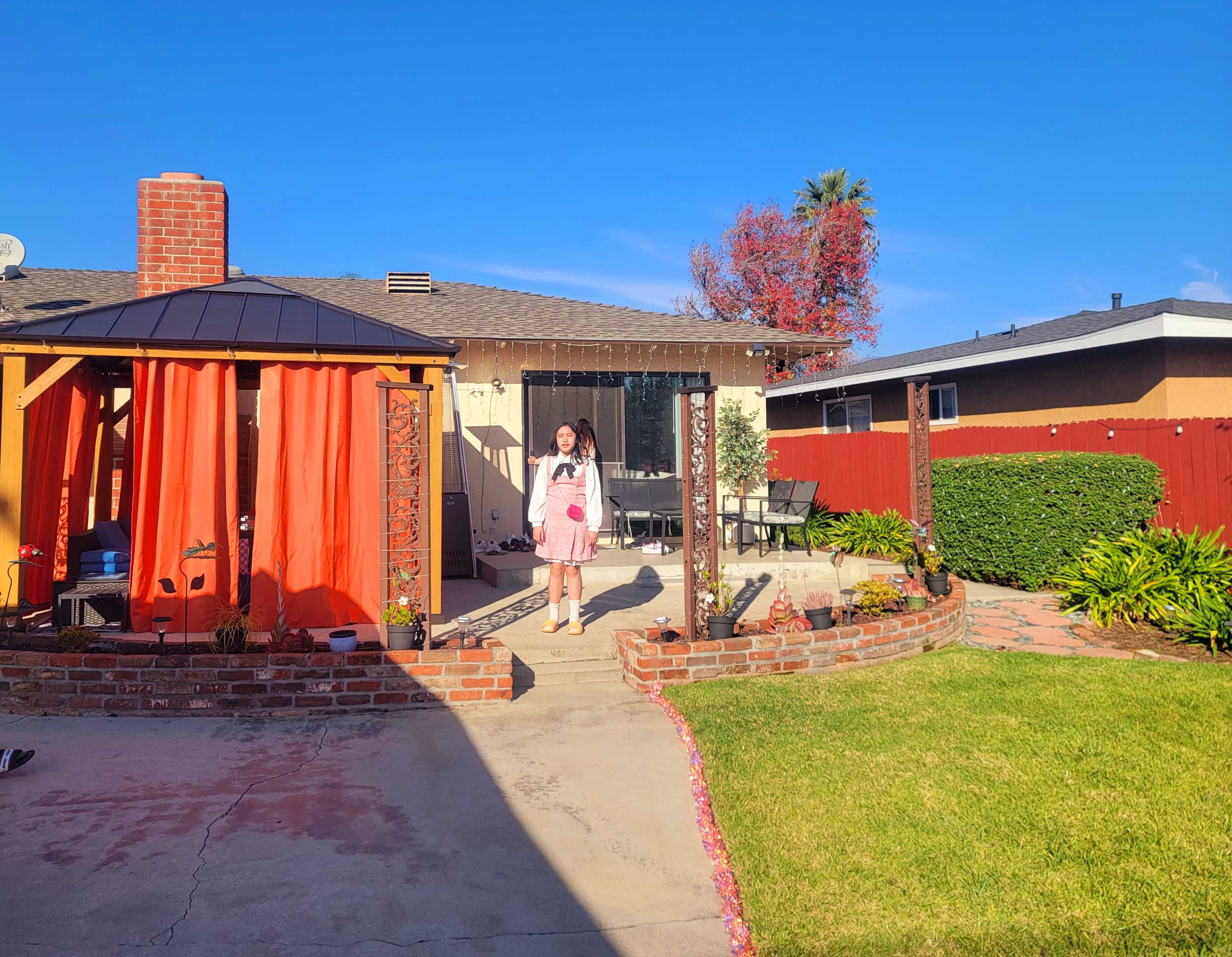 A woman stands in a backyard in front of a house with a brick border, a patio area, and colorful landscaping.