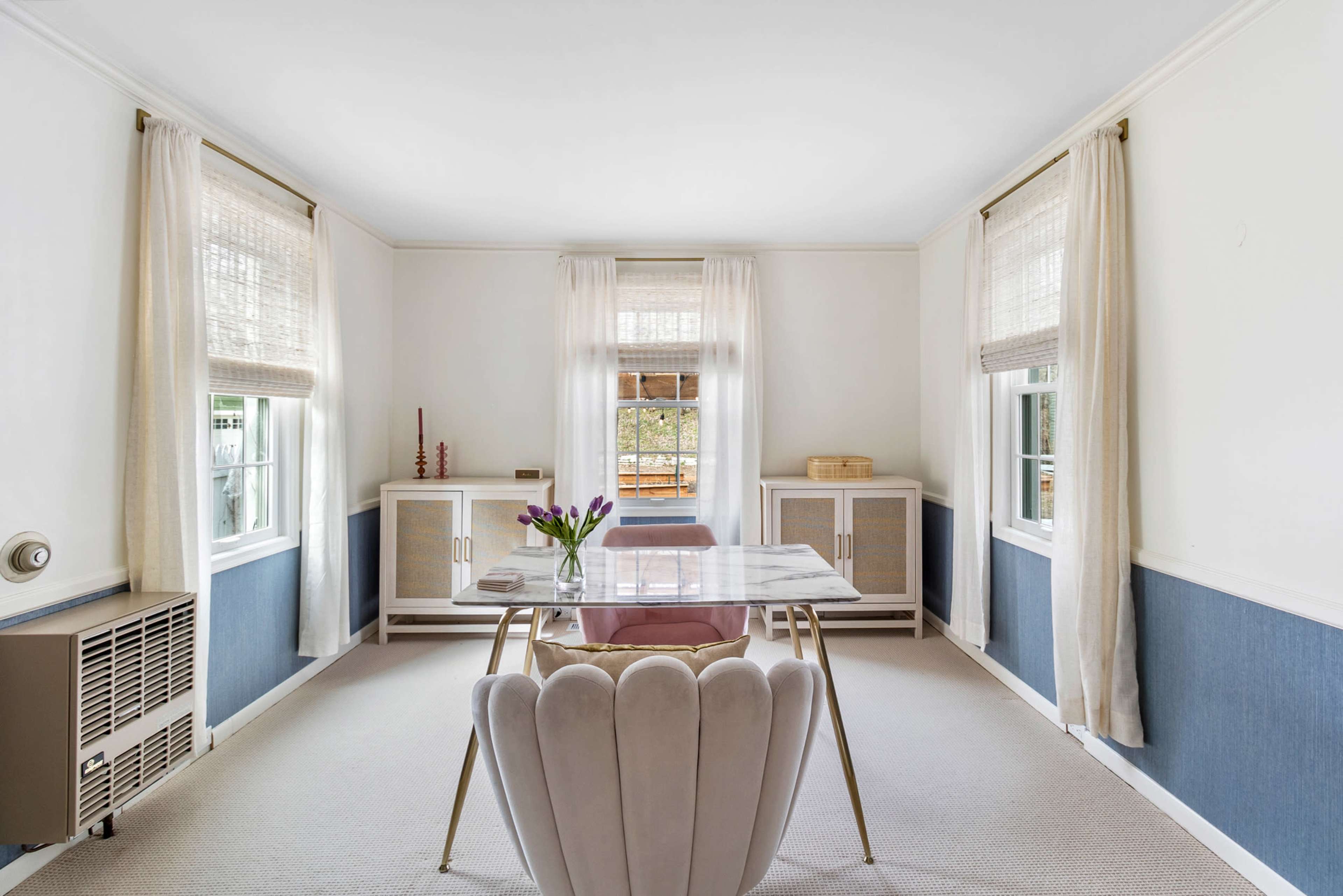 A well-lit office features a marble-top desk, a plush pink chair, and large windows with light curtains.