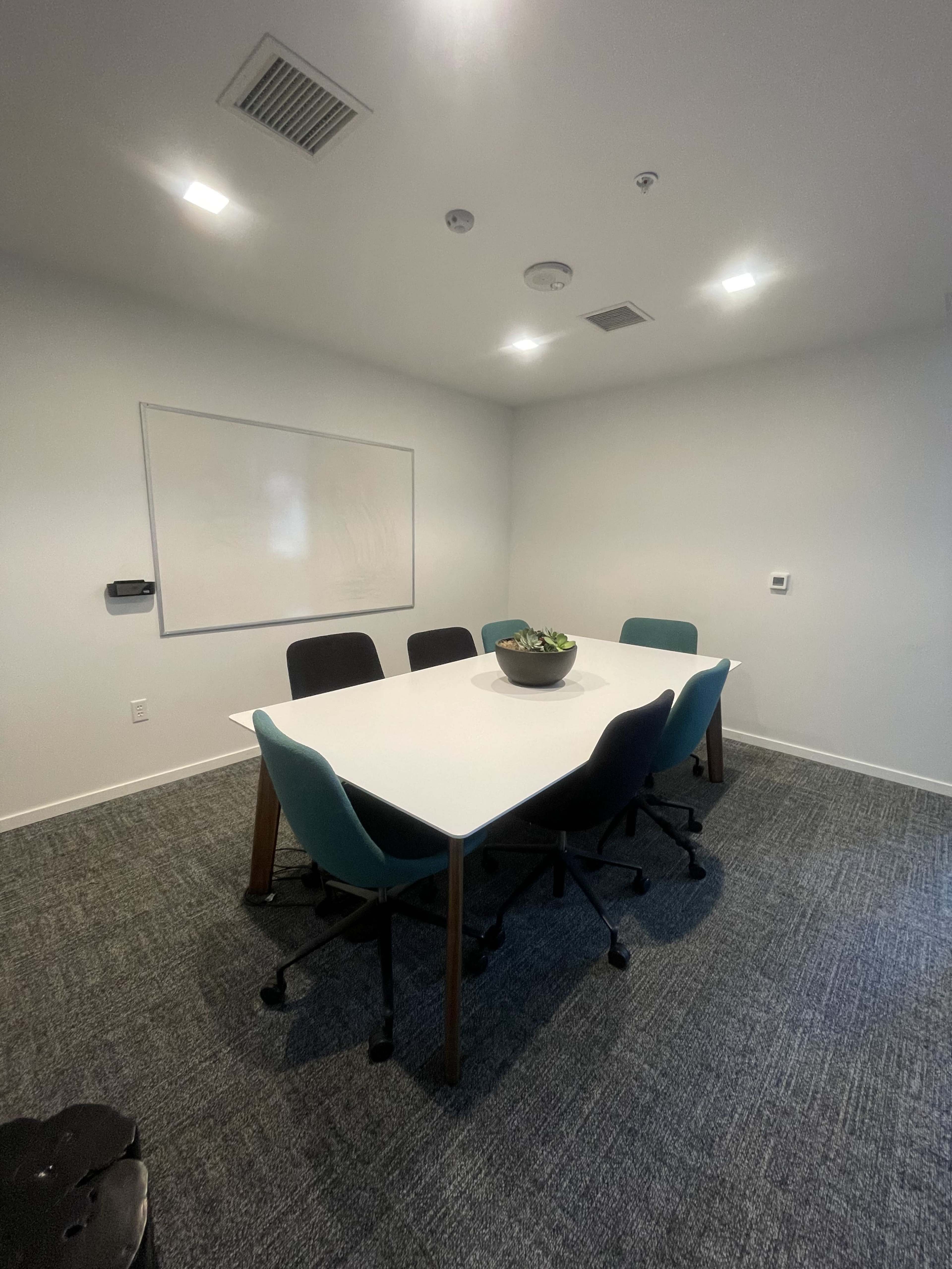 A conference room features a central table surrounded by six chairs, with a whiteboard on the wall and overhead lighting.